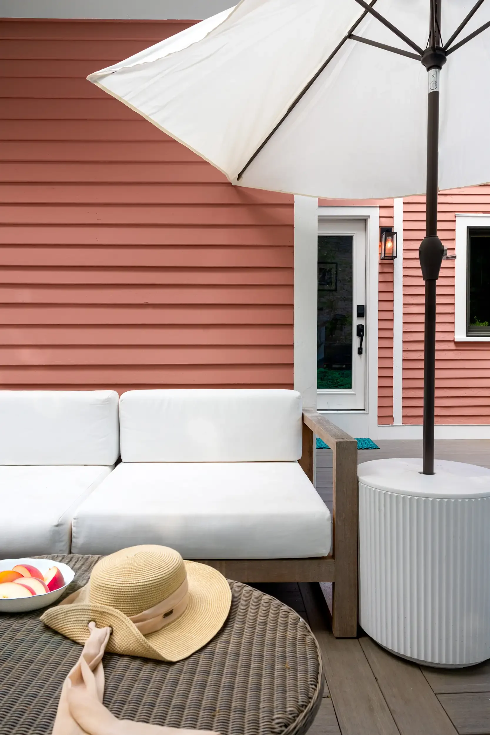 A backyard patio with a white outdoor sofa, a wicker table with a straw hat, sunhat, and bowl of apples, a large white patio umbrella, pink house siding, a glass door, a white window, and a small outdoor light fixture.