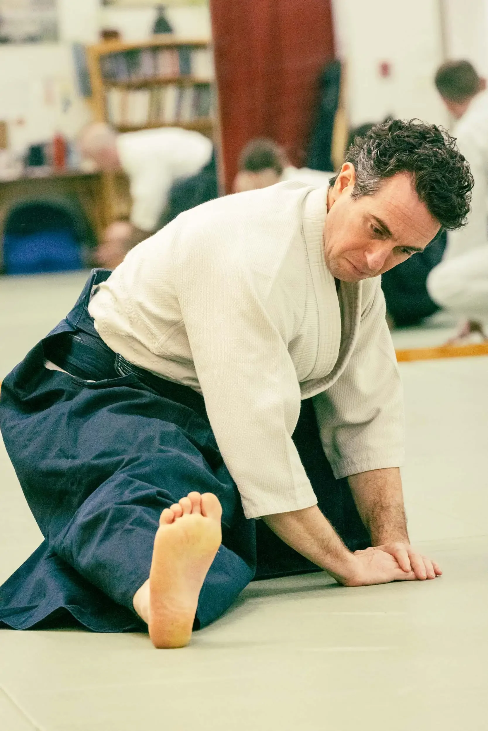 A man with curly dark hair wearing a beige kimono practicing martial arts on a mat with a child lying on their stomach. The child's face is not visible, and they are wearing dark blue pants. The setting appears to be a dojo or martial arts training r