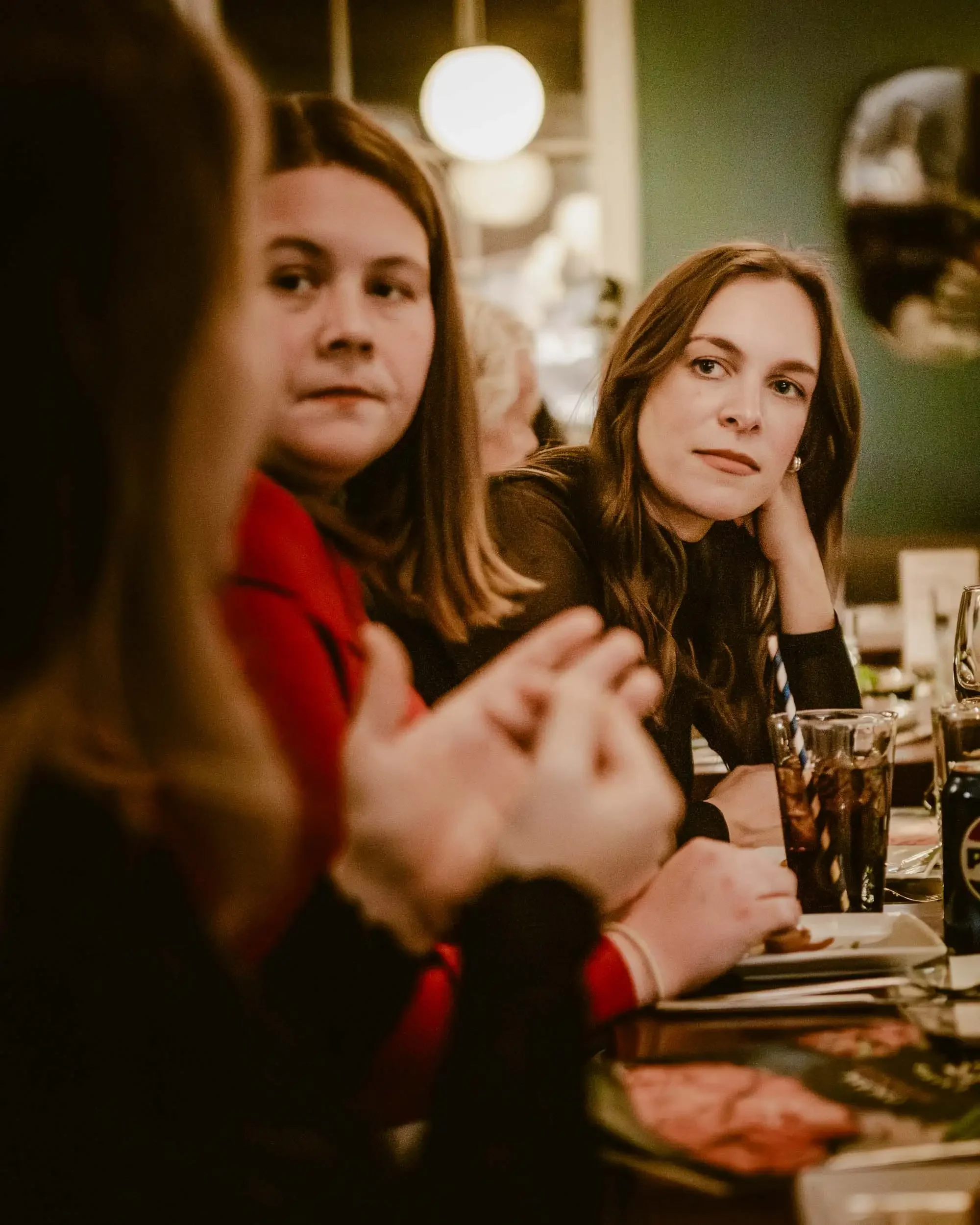 Three women seated at a restaurant table, two are clearly visible, one is partially visible, with drinks in front of them, and warm ambient lighting.