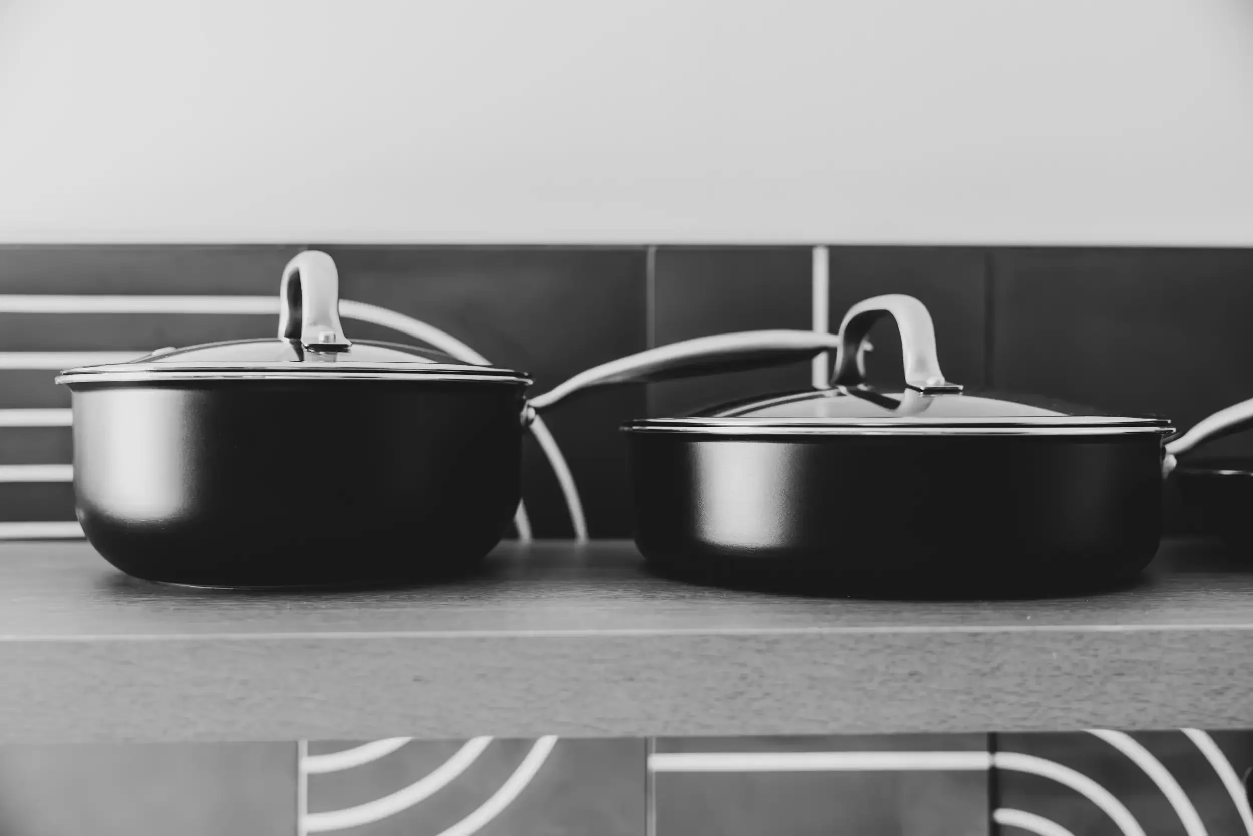Black and white photo of two stainless steel saucepans with lids on a kitchen stove. The background has horizontal tile and stove burners visible.