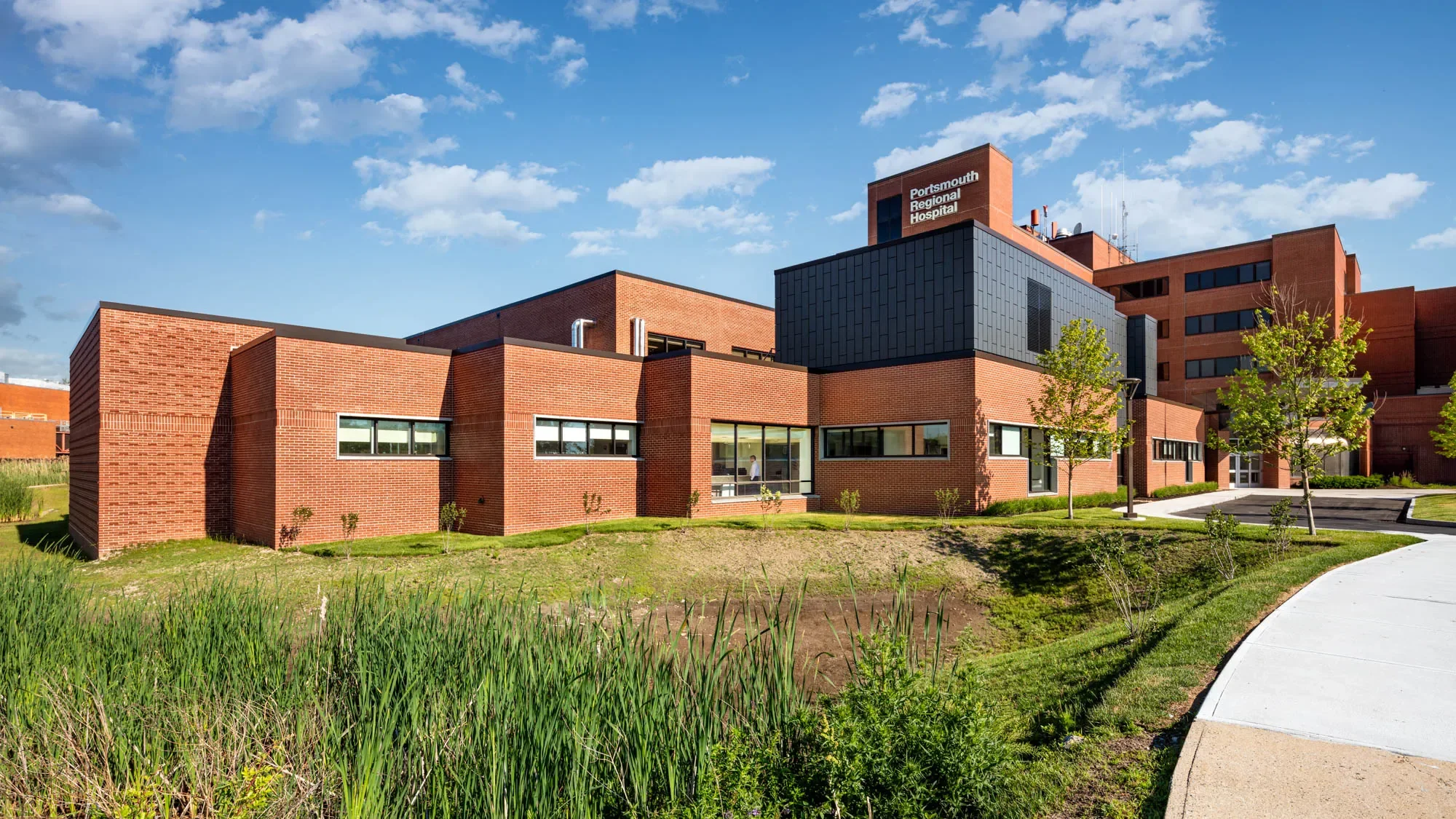 Exterior view of Portsmouth Regional Hospital building with brick walls, black and gray accents, and a blue sky with scattered clouds.