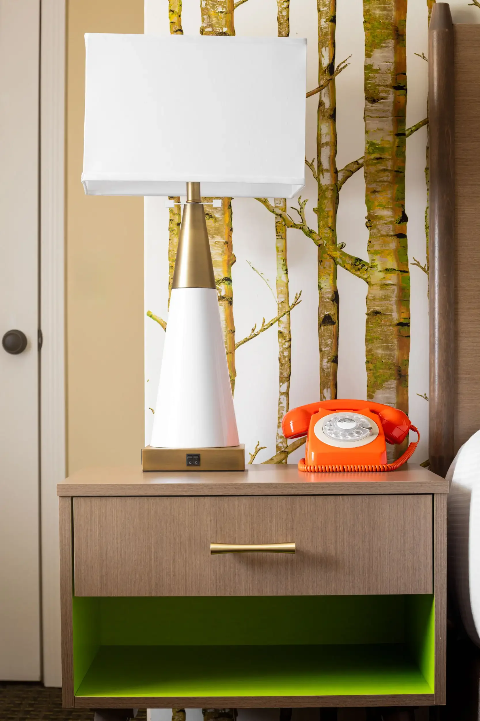 A wooden nightstand with a beige finish and a gold handle, featuring an open space with a lime green interior. On top of the nightstand, there is a retro orange rotary telephone and a modern white table lamp with a conical base and a rectangular whit
