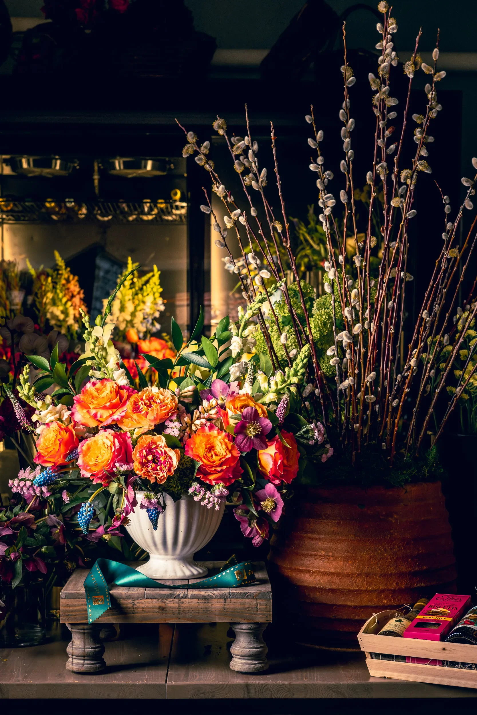 Arrangement of multicolored flowers in white and terracotta pots on wooden tables with ribbons and a box of chocolates nearby.