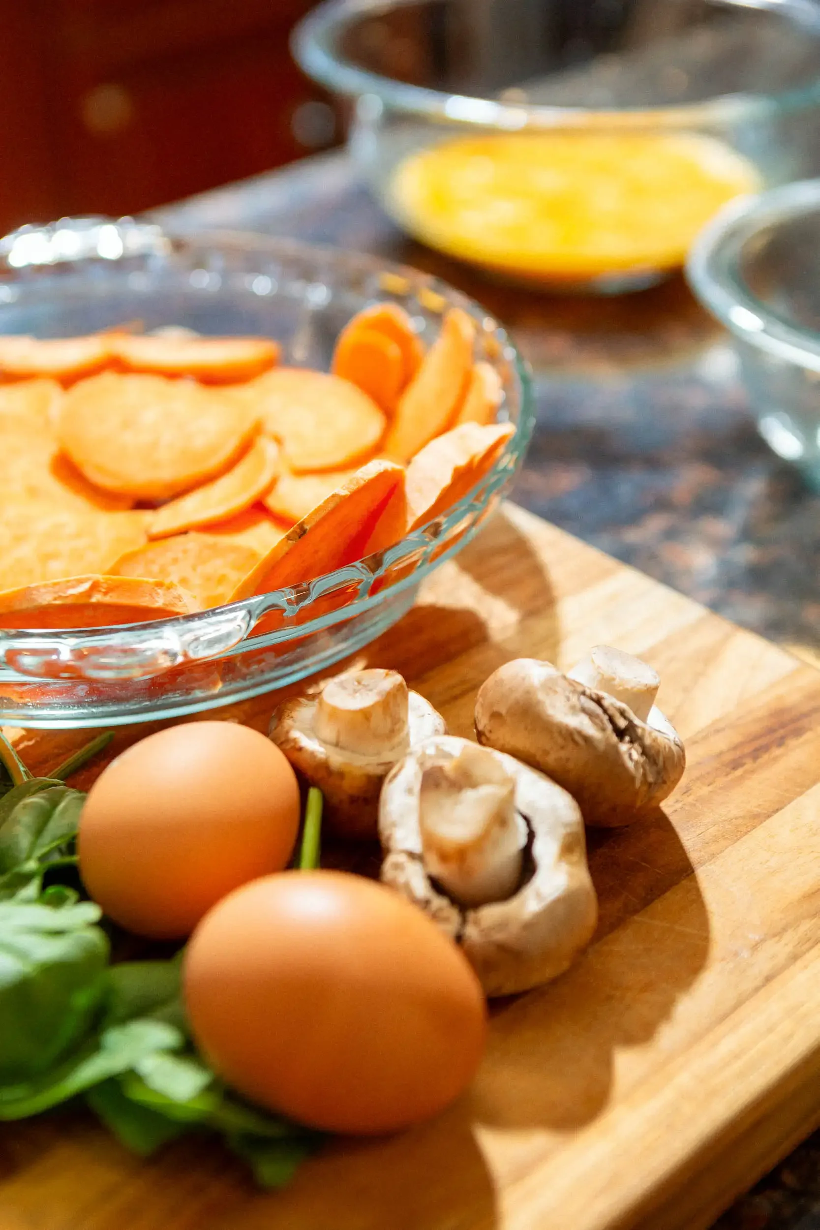 Eggs, mushrooms, and greens on a wooden cutting board, with bowls of sliced vegetables in the background.