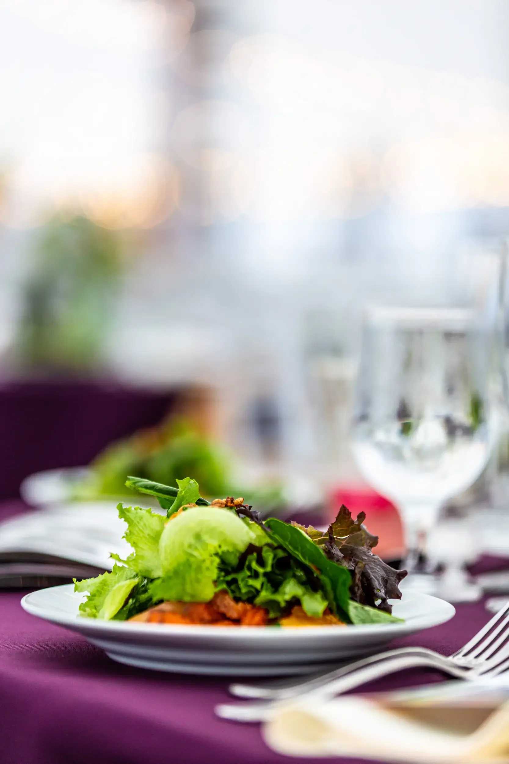A plate of salad with green and purple leafy lettuce, tomato, and other vegetables on a white dish, set on a purple tablecloth with a fork and knife beside it. In the background, there are blurred glasses and a window with light coming through.