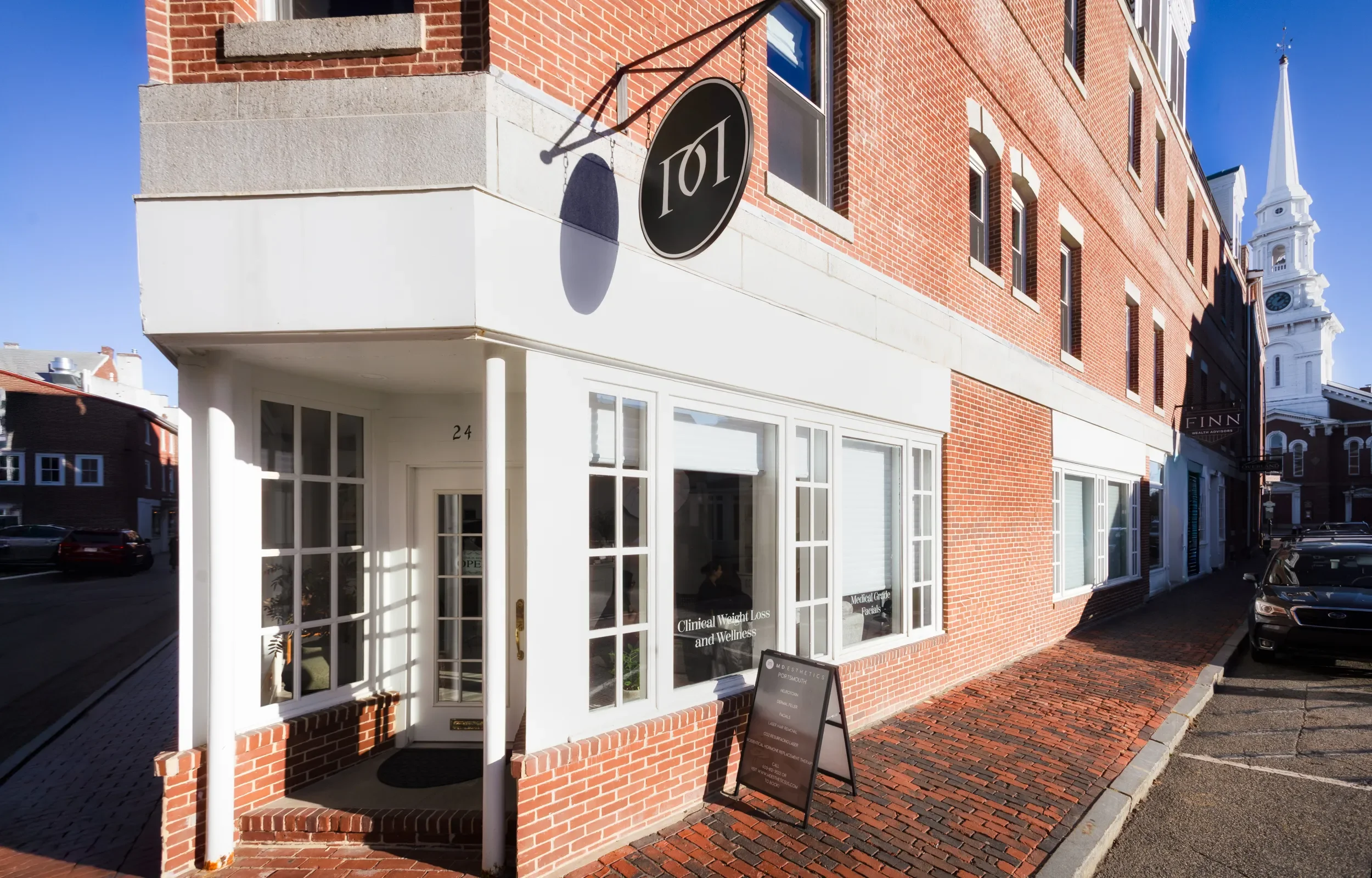 Storefront with a sign that reads '101' and large glass windows displaying clinical weight loss and wellness services, located in a brick building with a white entrance and a black sandwich board sign outside.