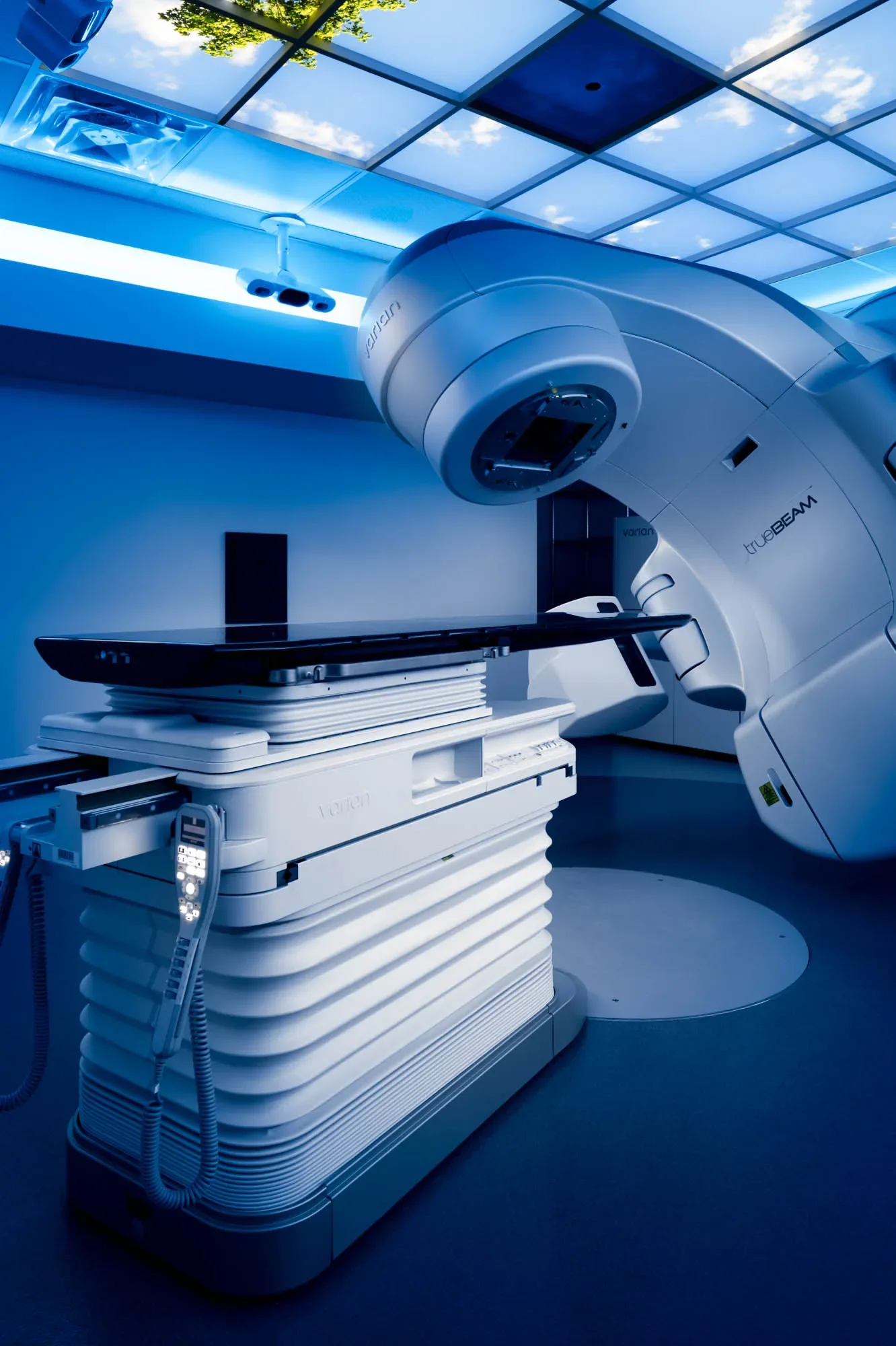 A medical room featuring a large, modern MRI machine with a sleek, white design, situated next to a table for patient positioning. The room has a blue lighting ambiance and a ceiling with reflective panels showing a cloudy sky and green trees outside
