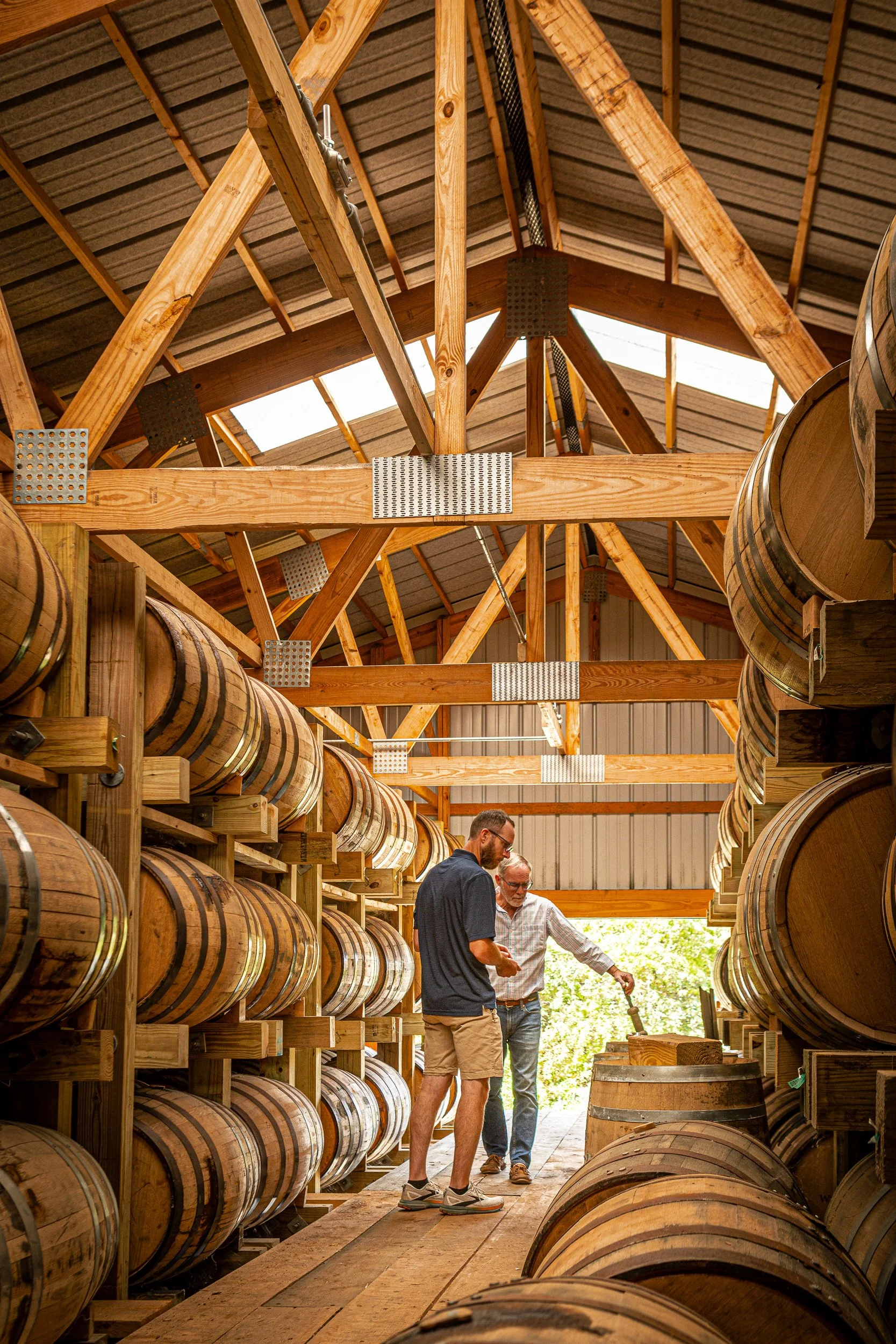 Two men in a barrel aging cellar, inspecting wooden barrels used for aging spirits or wine, with a wooden ceiling overhead.