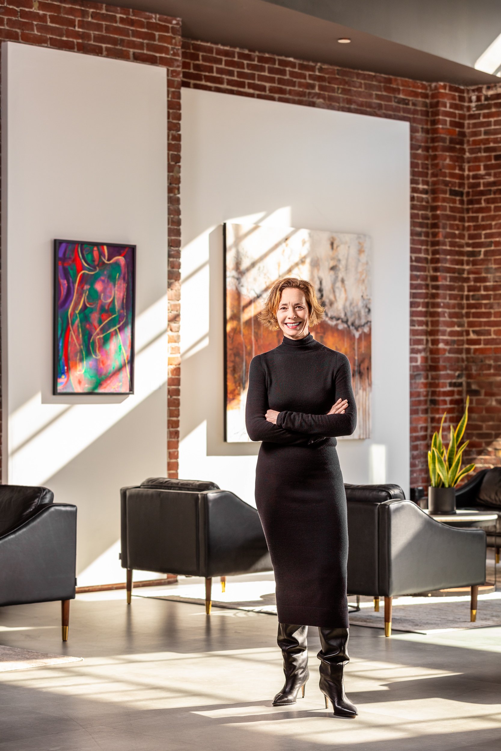 A woman with short curly hair wearing a black turtleneck dress and black thigh-high boots standing with arms crossed in an art gallery, with abstract paintings and brick walls in the background.