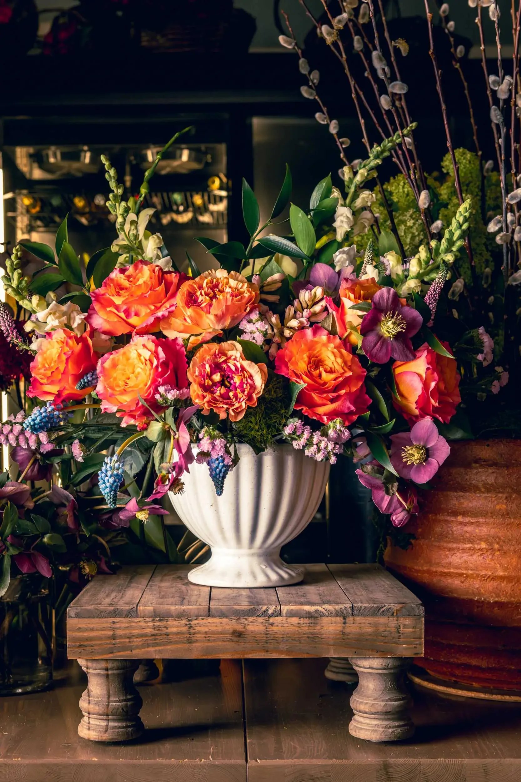 A large floral arrangement in a white ceramic vase with a fluted design and pedestal base, placed on a rustic wooden table. The arrangement contains orange and pink peonies, white snapdragons, purple hellebores, green leaves, and tall pussy willow br