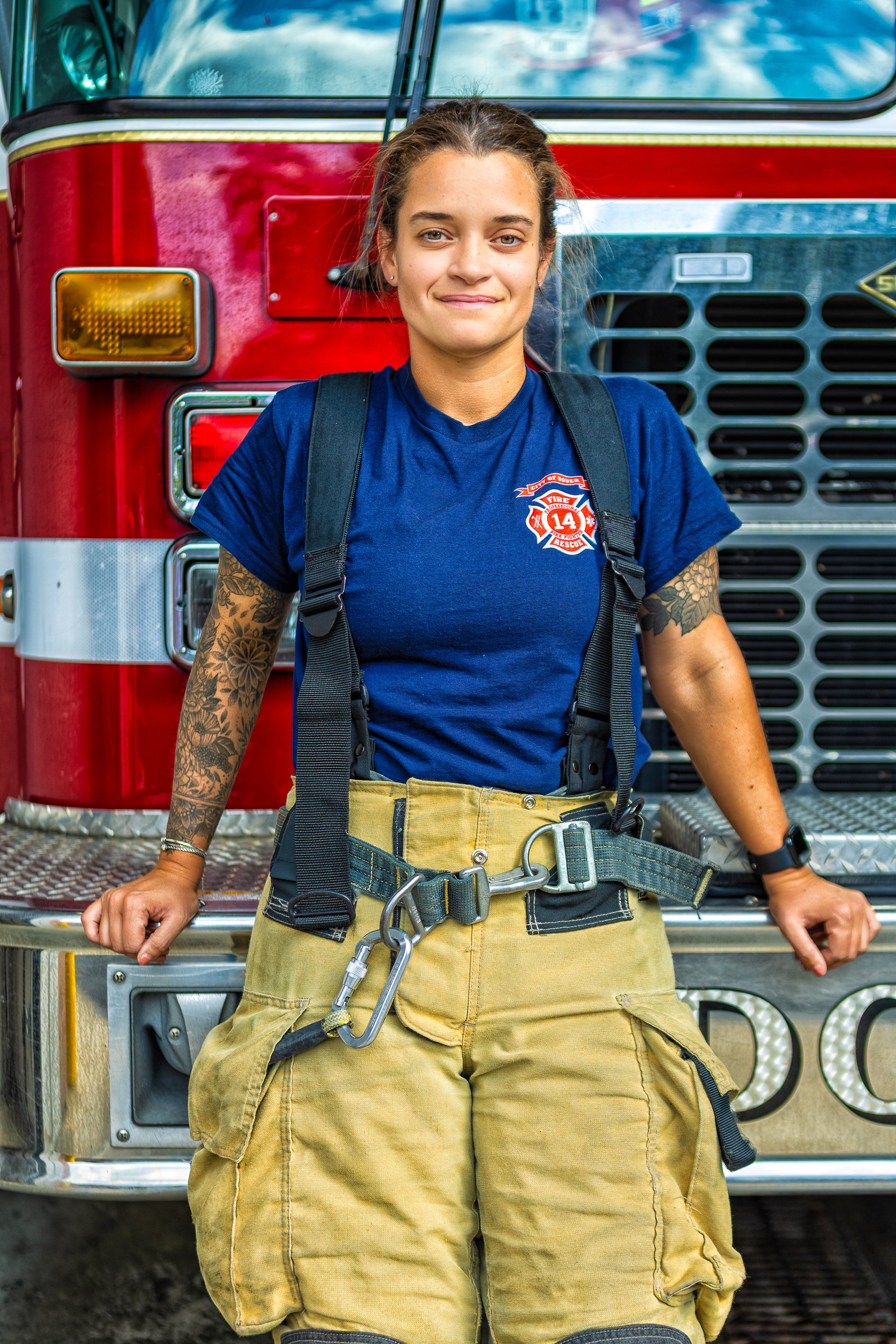 A young female firefighter standing in front of a fire truck, wearing a blue shirt with a fire department badge, firefighter's pants, and safety gear.
