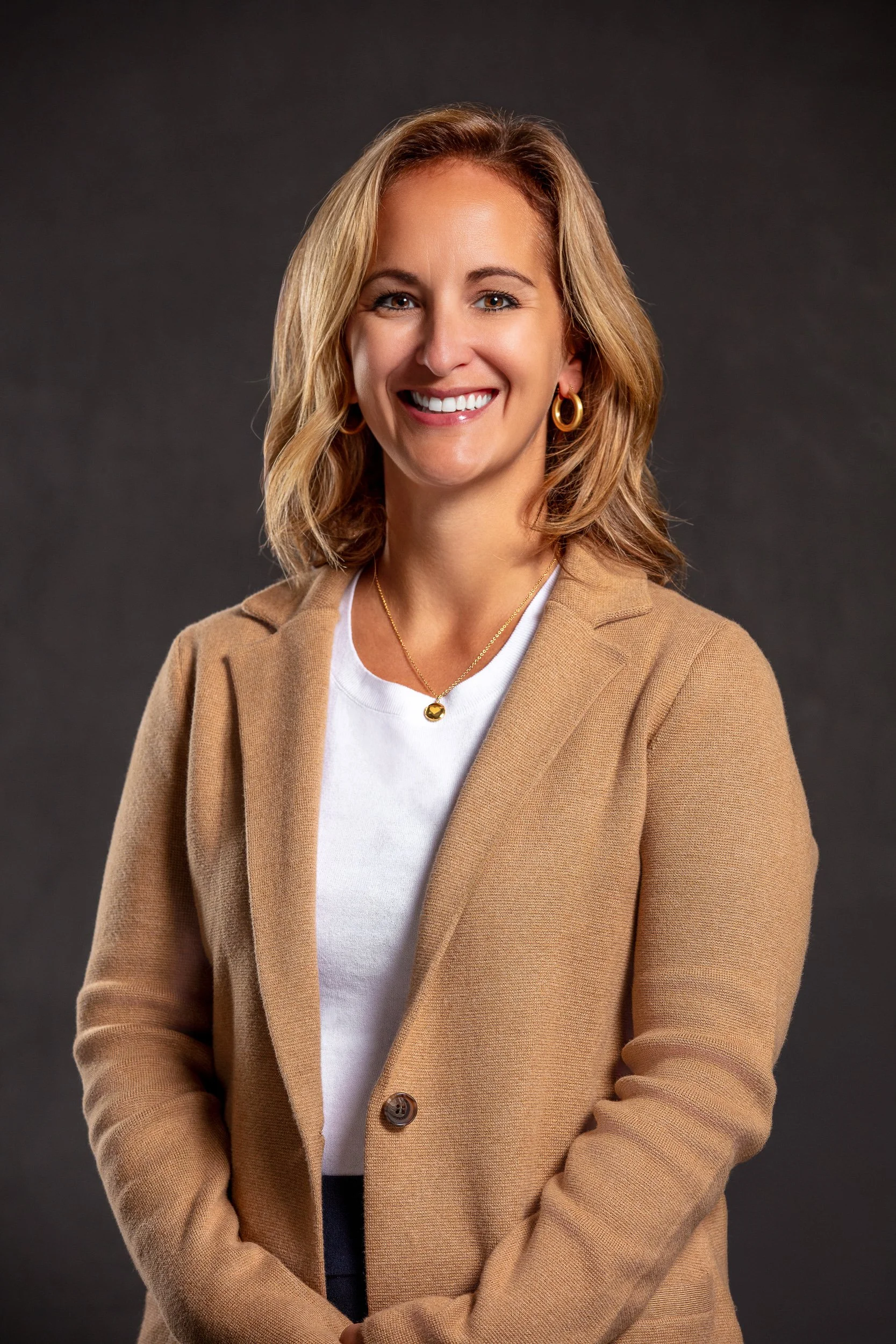 A woman with shoulder-length blonde hair, wearing a beige blazer, white t-shirt, gold jewelry, and smiling at the camera against a dark gray background.