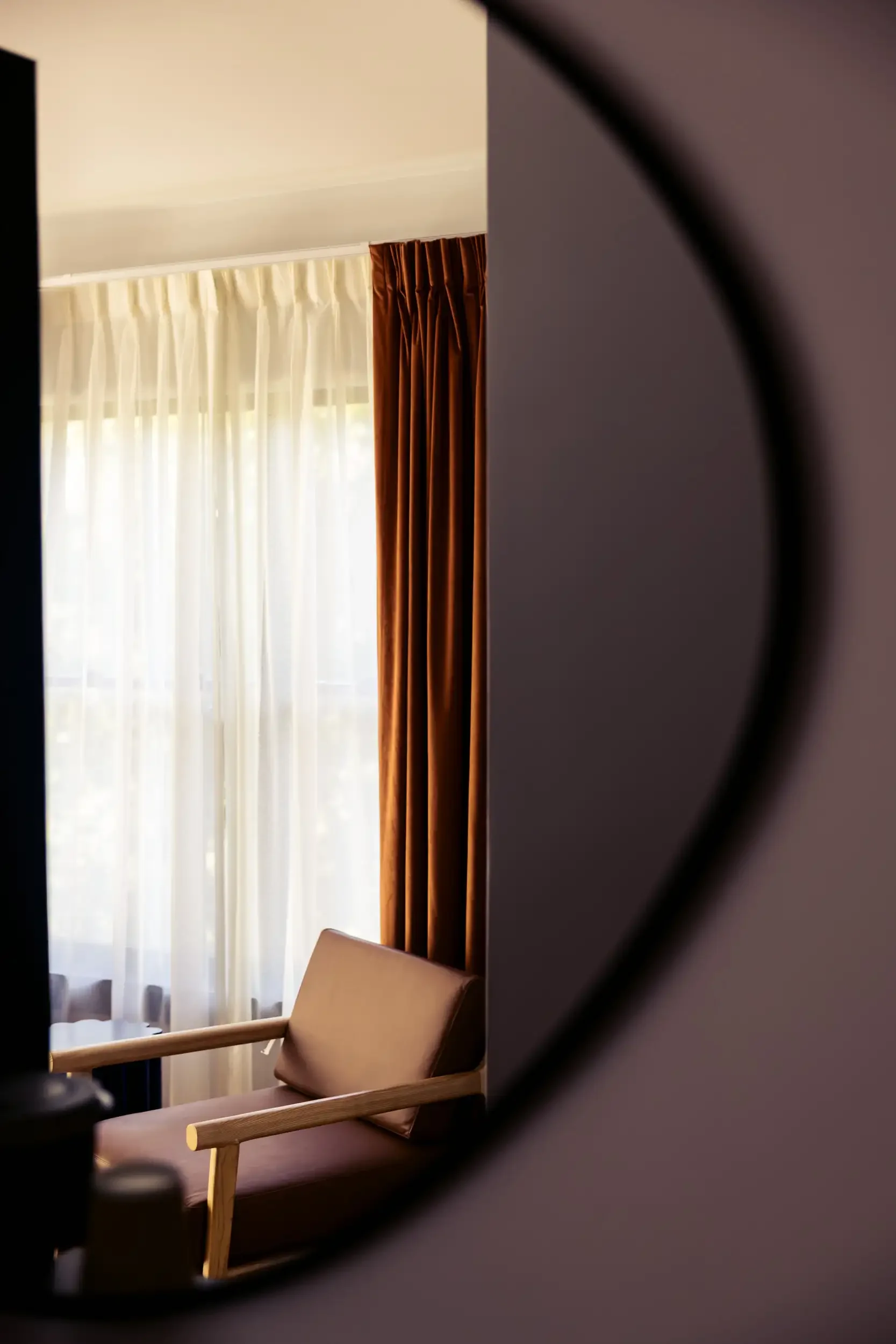 A cozy corner of a room with beige and brown curtains, a beige armchair with a cushion, and sunlight filtering through sheer curtains.