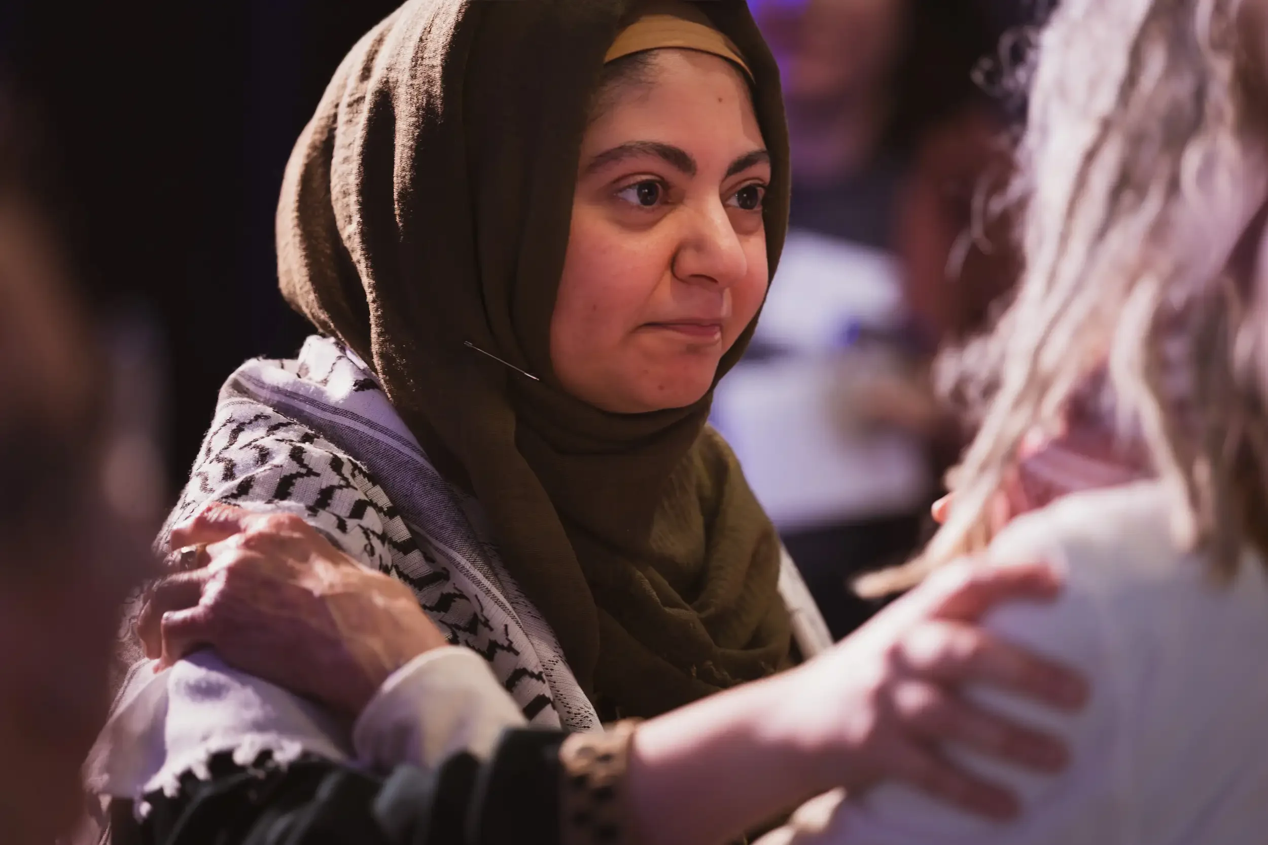 Close-up of a woman with a brown headscarf and a patterned outer garment, looking intently at someone or something, with soft lighting and blurred background.