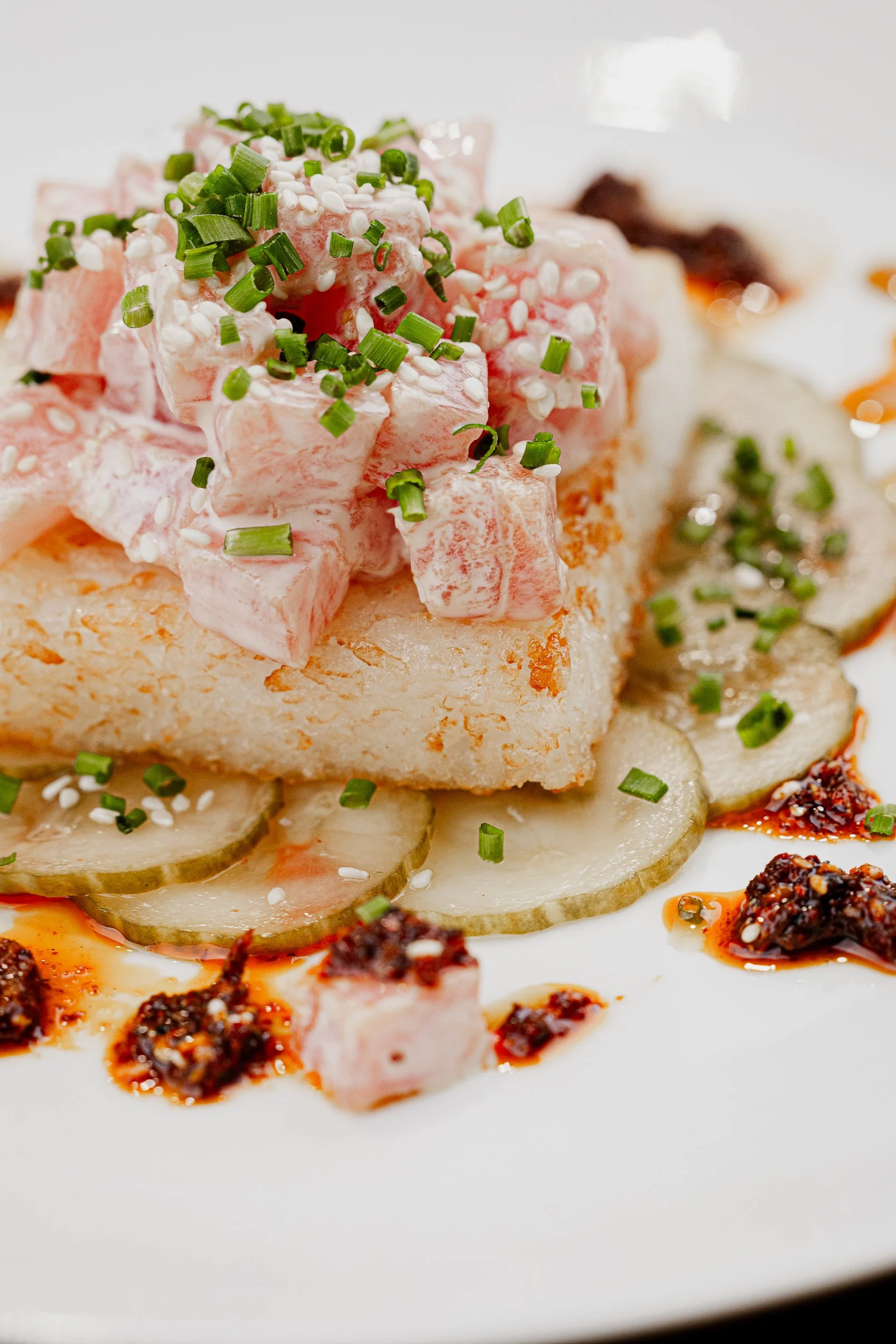 Close-up of a gourmet dish with diced ham, chopped green onions, sliced cucumbers, and sauces on a white plate.