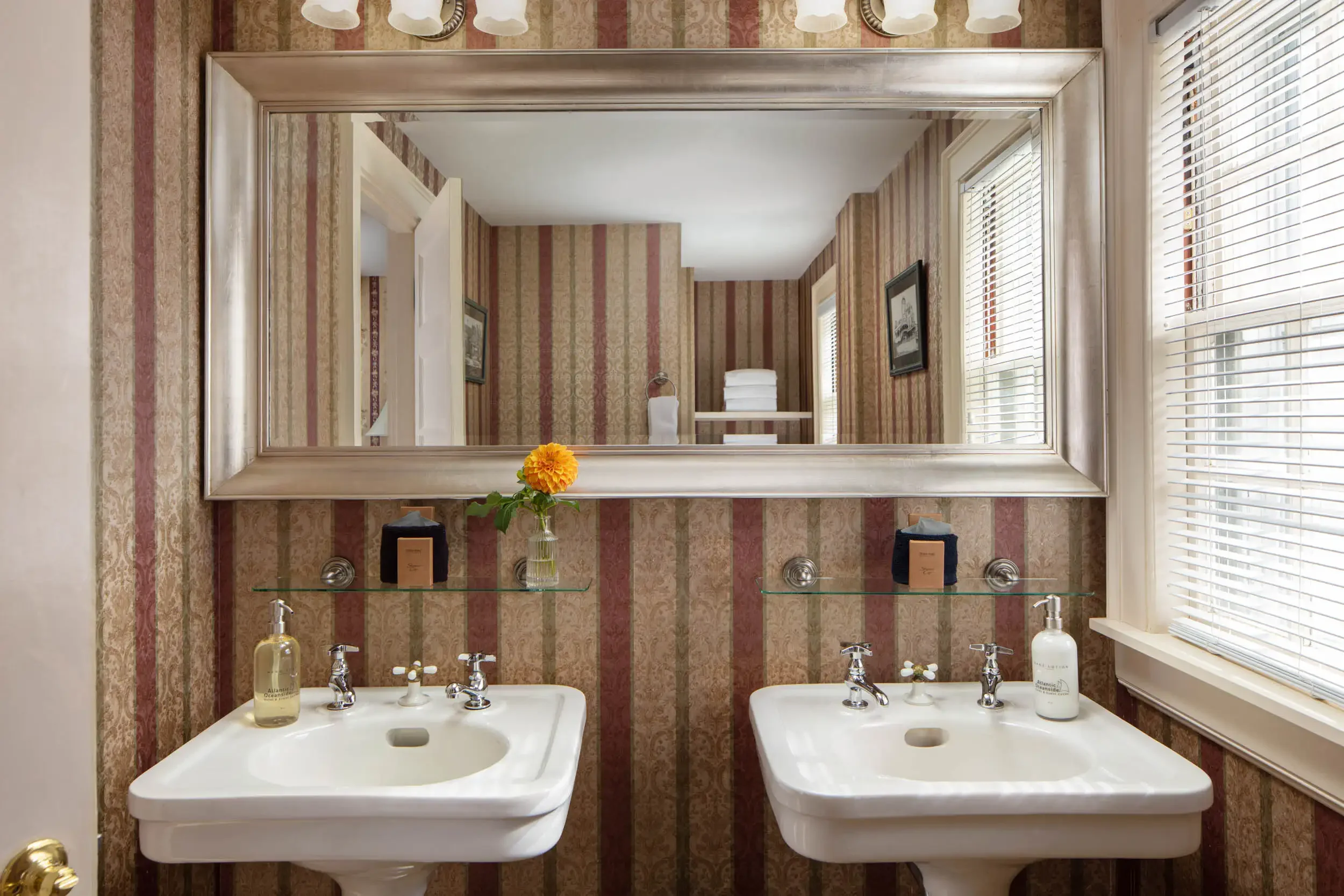 Double bathroom sinks with liquid soap bottles and a large mirror above. There is a window with wooden blinds to the right and wallpaper with vertical stripes in beige and red tones. A small table in front of the mirror holds a glass vase with an ora