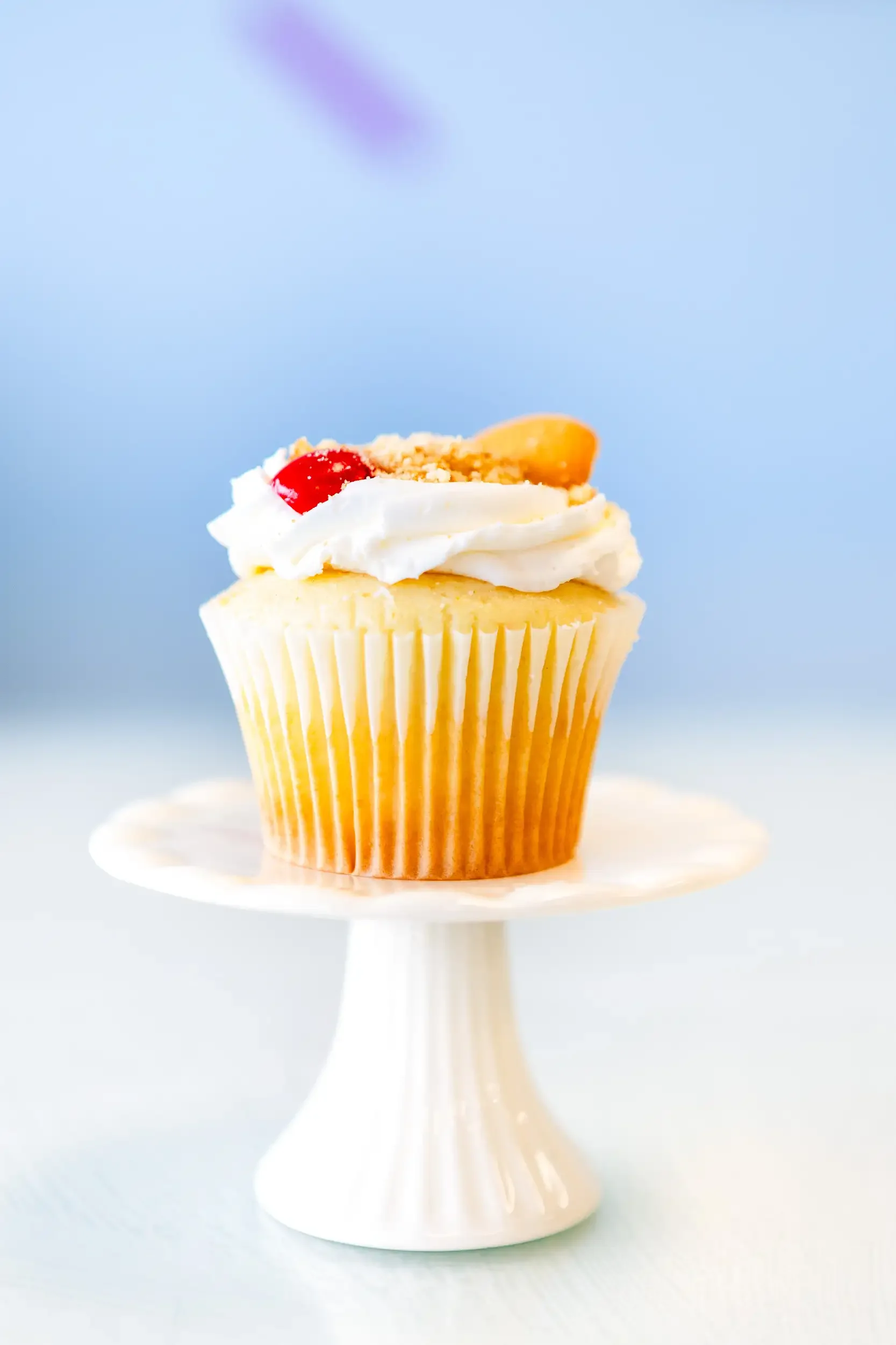 A cupcake with white frosting topped with a cherry and crunch crumbs, displayed on a white cake stand with a light blue background.