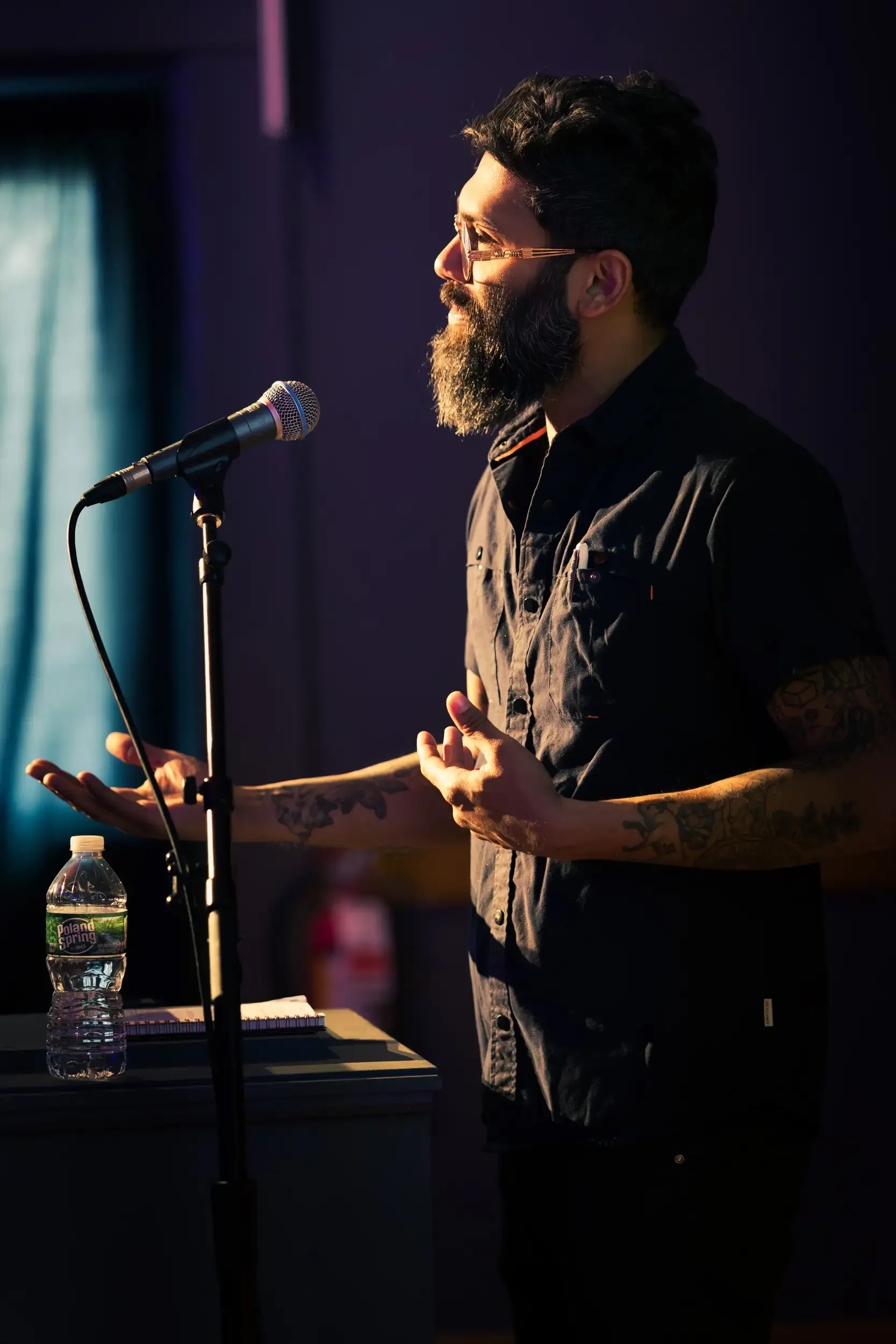 A man with dark hair, glasses, and a beard is speaking into a microphone. He has tattoos on his arms and is wearing a black shirt. On a table in front of him, there is a water bottle, a notebook, and a pen. The background is dark with some lighting o