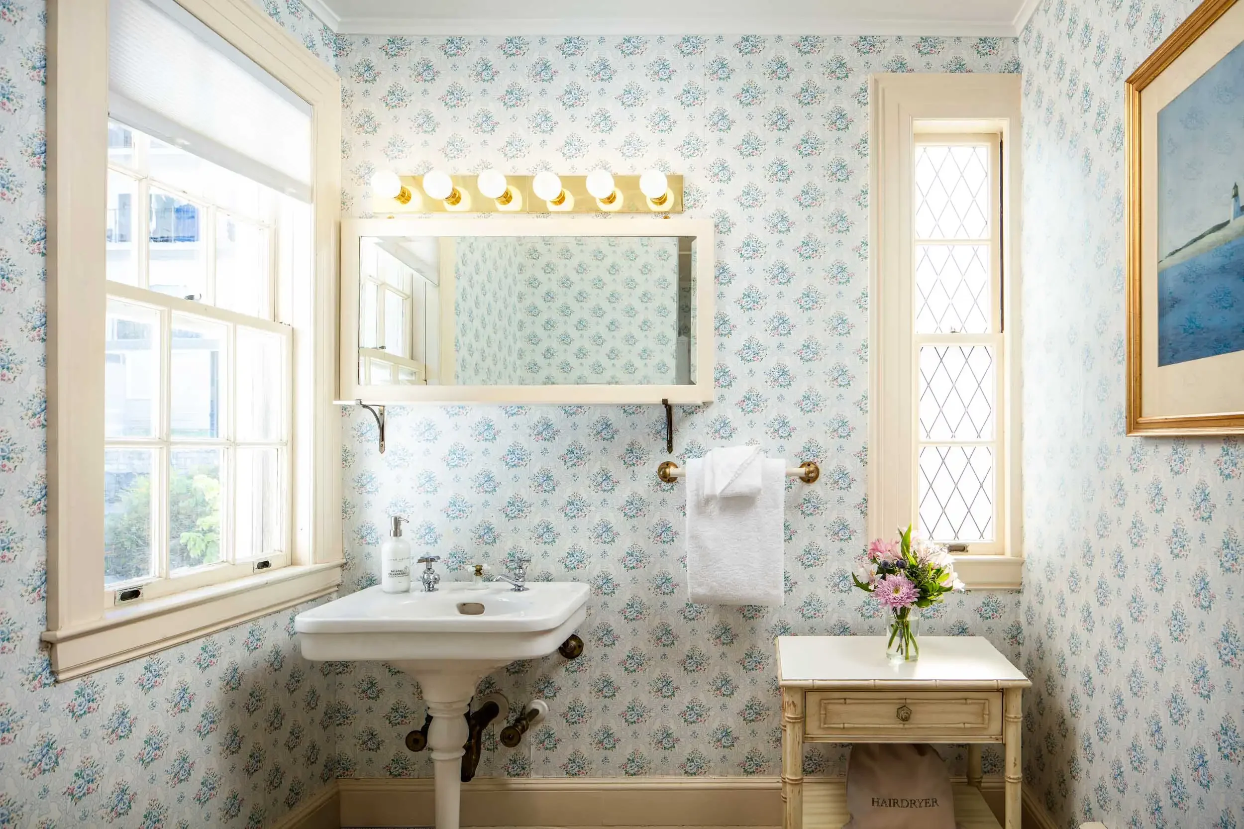 A vintage bathroom with floral wallpaper, a white pedestal sink, a mirror, and a small wooden table with a vase of pink and purple flowers, with natural light coming through two windows.
