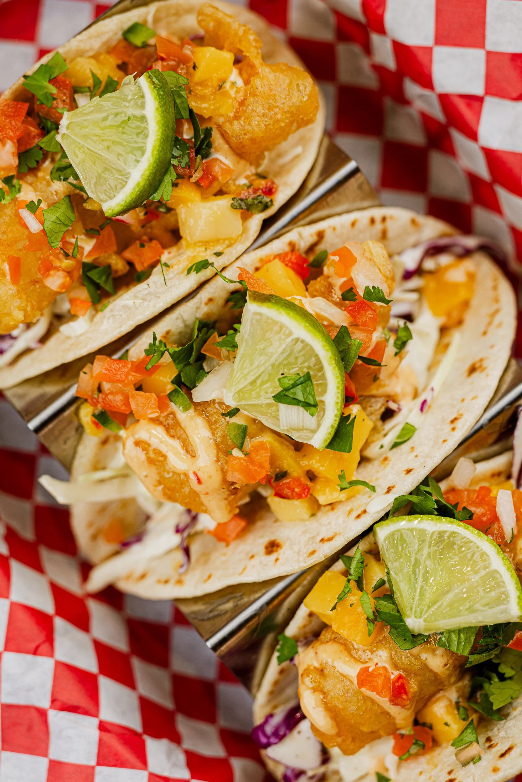 Three fish tacos topped with lime wedges, chopped vegetables, and cilantro, served on a basket lined with red and white checkered paper.
