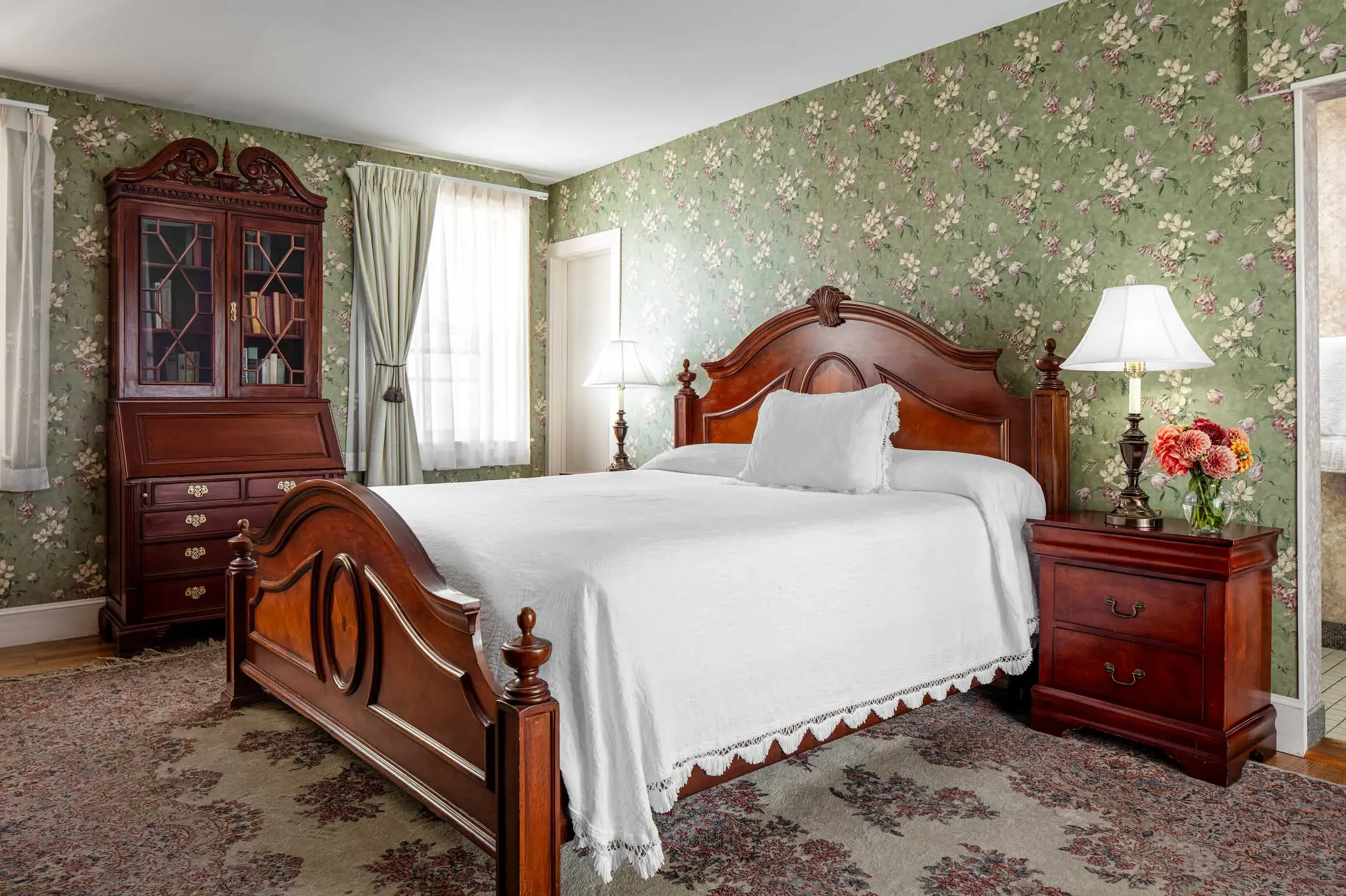 A bedroom with green floral wallpaper, a wooden bed with white bedding and a pillow, two matching wooden nightstands with lamps, a tall wooden cabinet with glass doors, and curtains on the window.