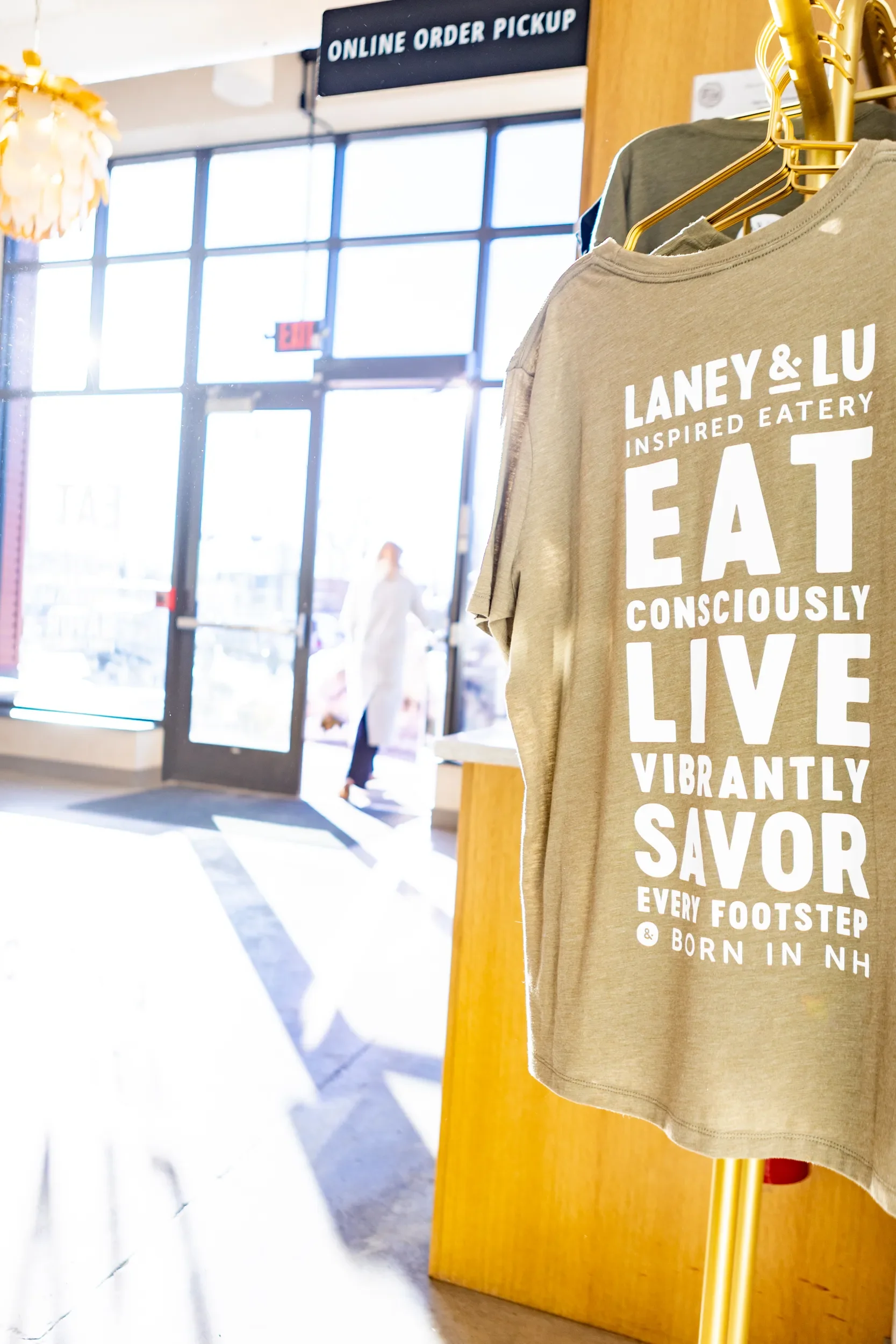 A beige T-shirt on display in a store with white bold text promoting conscious eating and vibrant living. In the background, there's a store entrance labeled 'Online Order Pickup,' with sunlight streaming through large windows.
