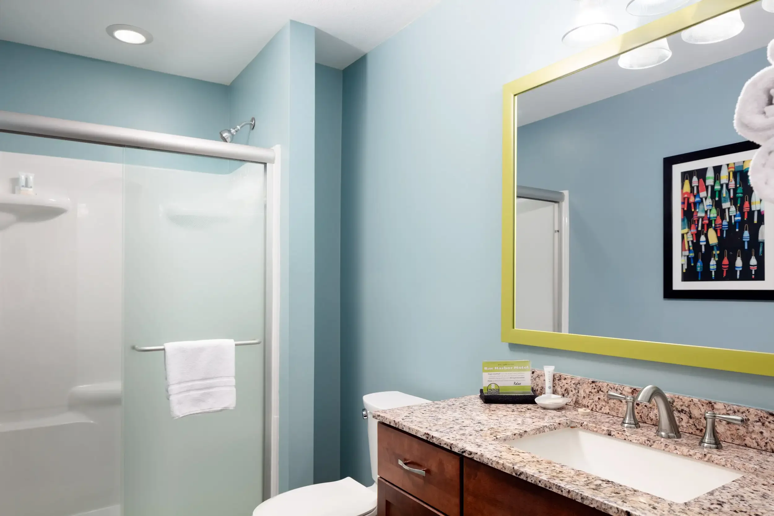 A bathroom with a granite countertop sink, a large mirror with a yellow frame, a shower with a glass door, and a wall art of colorful popsicles.