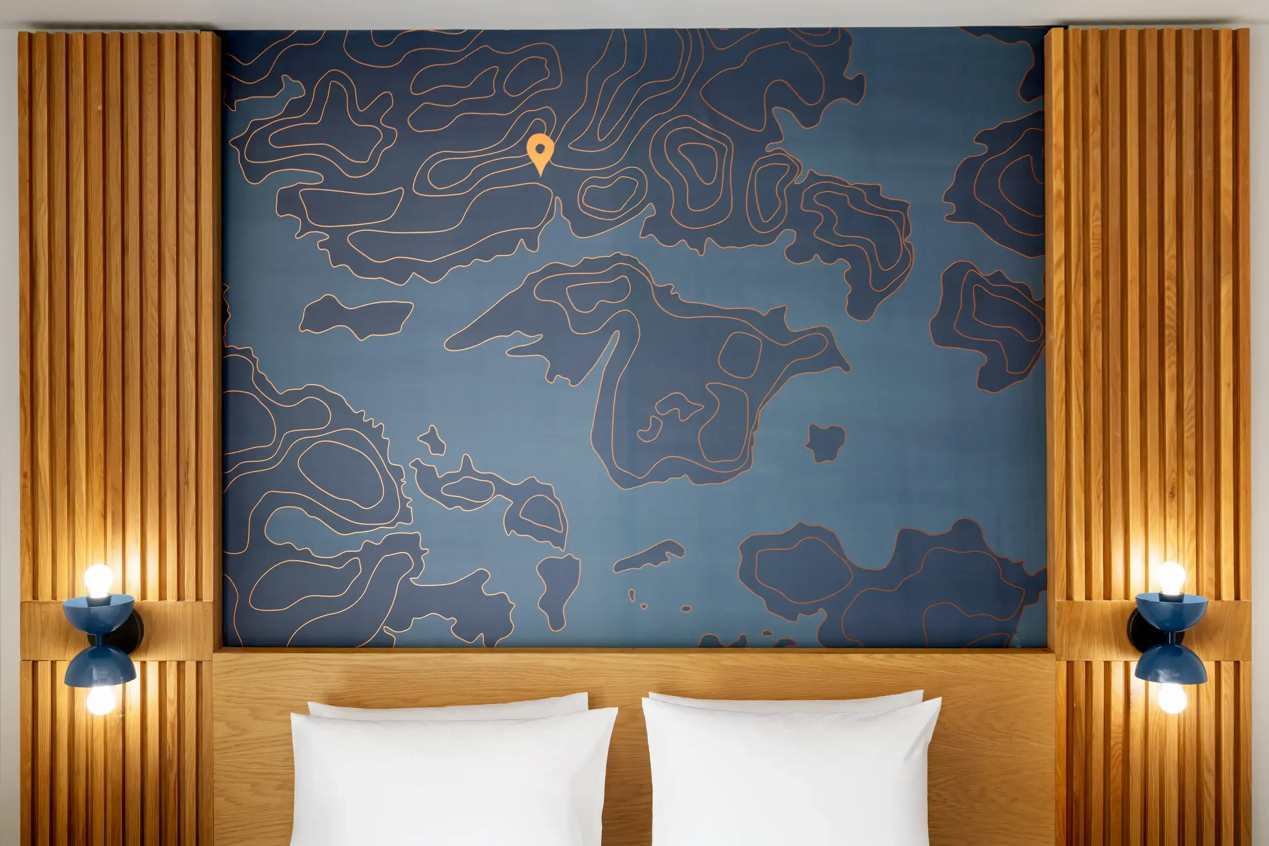 A bedroom with a large wall art piece featuring a topographical map behind a bed with white pillows, flanked by wooden slat wall panels, and two blue bedside lamps with exposed light bulbs.