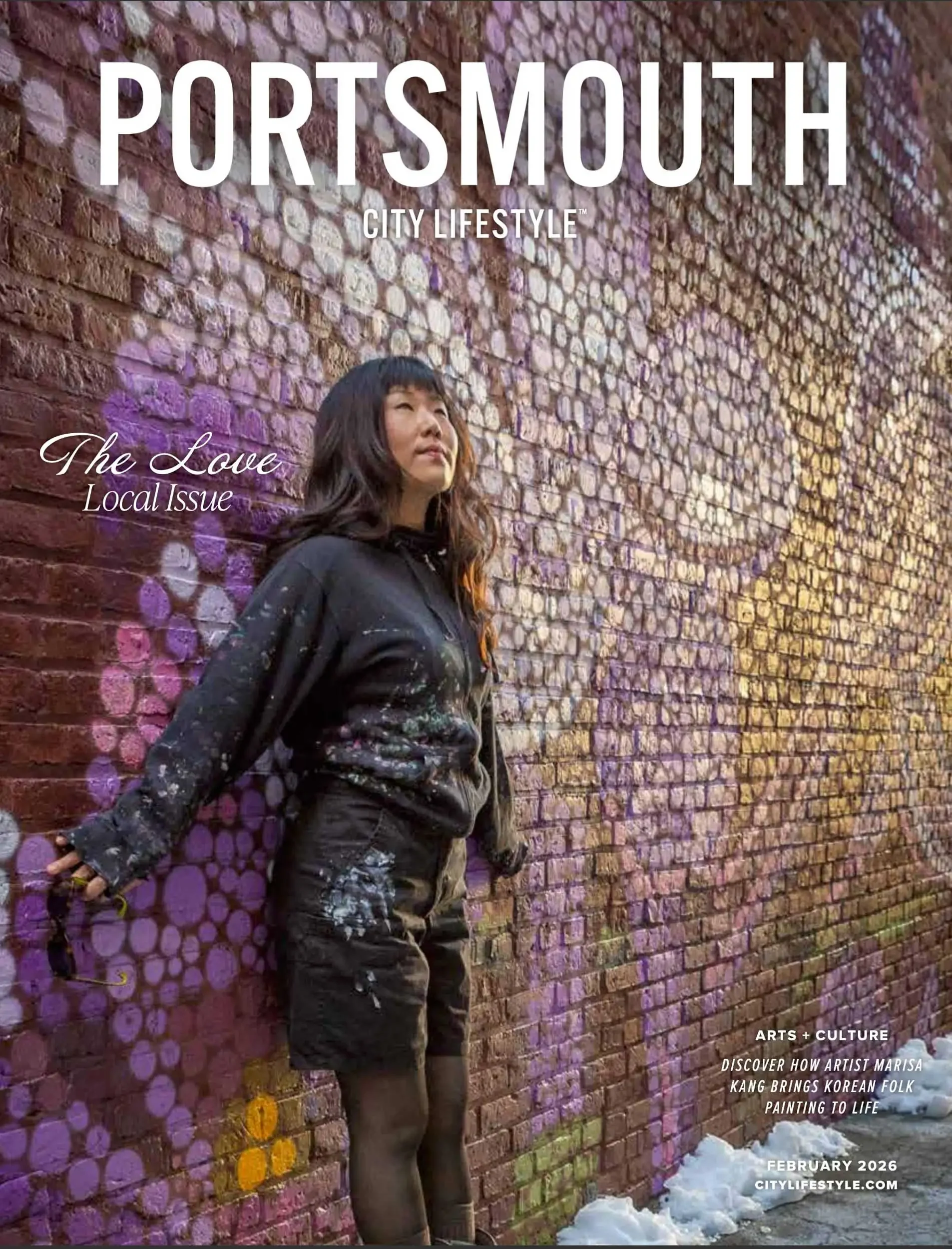 A woman stands against a brick wall with purple and yellow circular patterns painted on it. She is wearing a black jacket and black pants with paint splatters, with snow on the ground nearby. The image features text promoting a cultural event in Port
