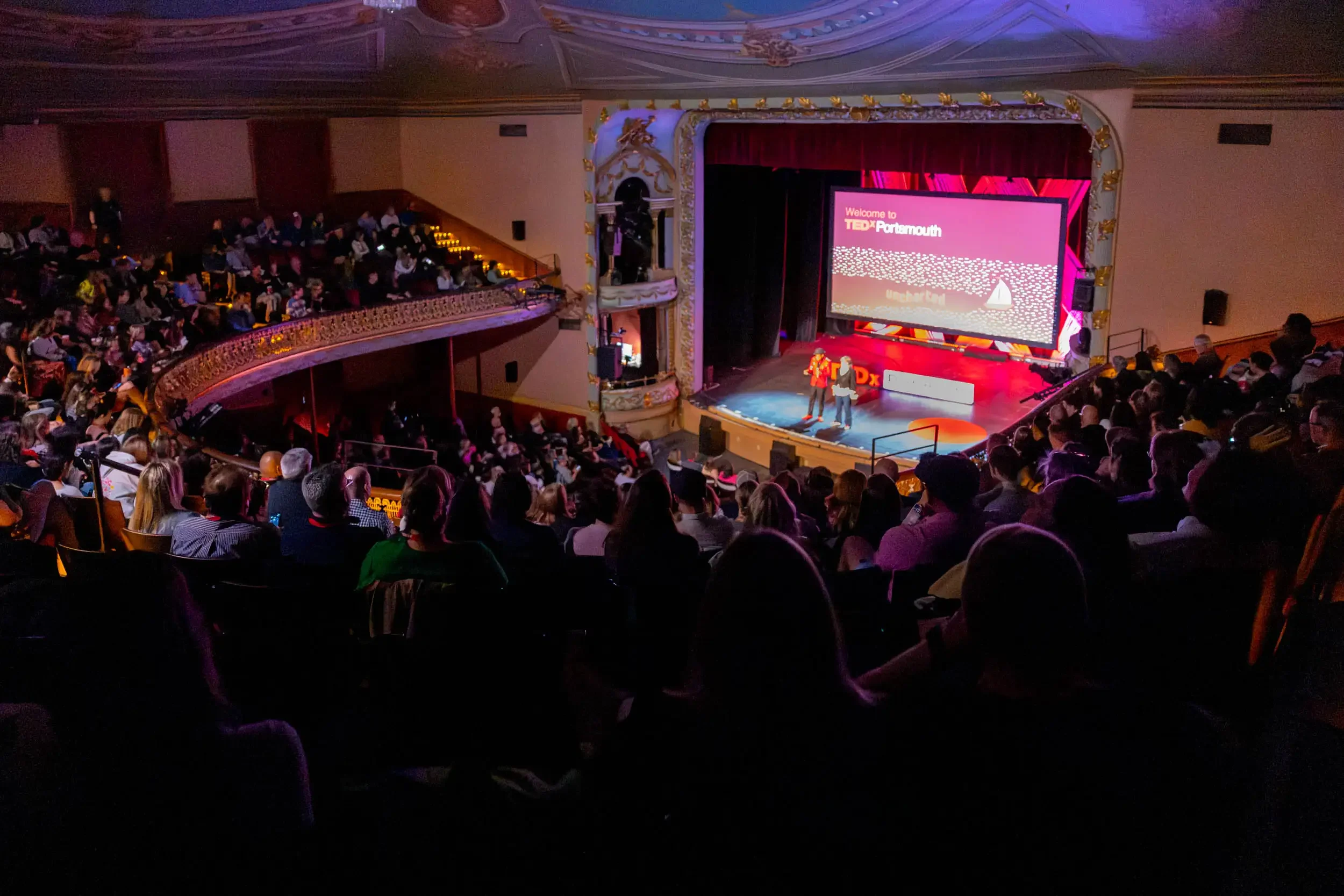 Audience seated in theater watching a TEDxPortsmouth event on stage with a large digital screen and two speakers standing in front.