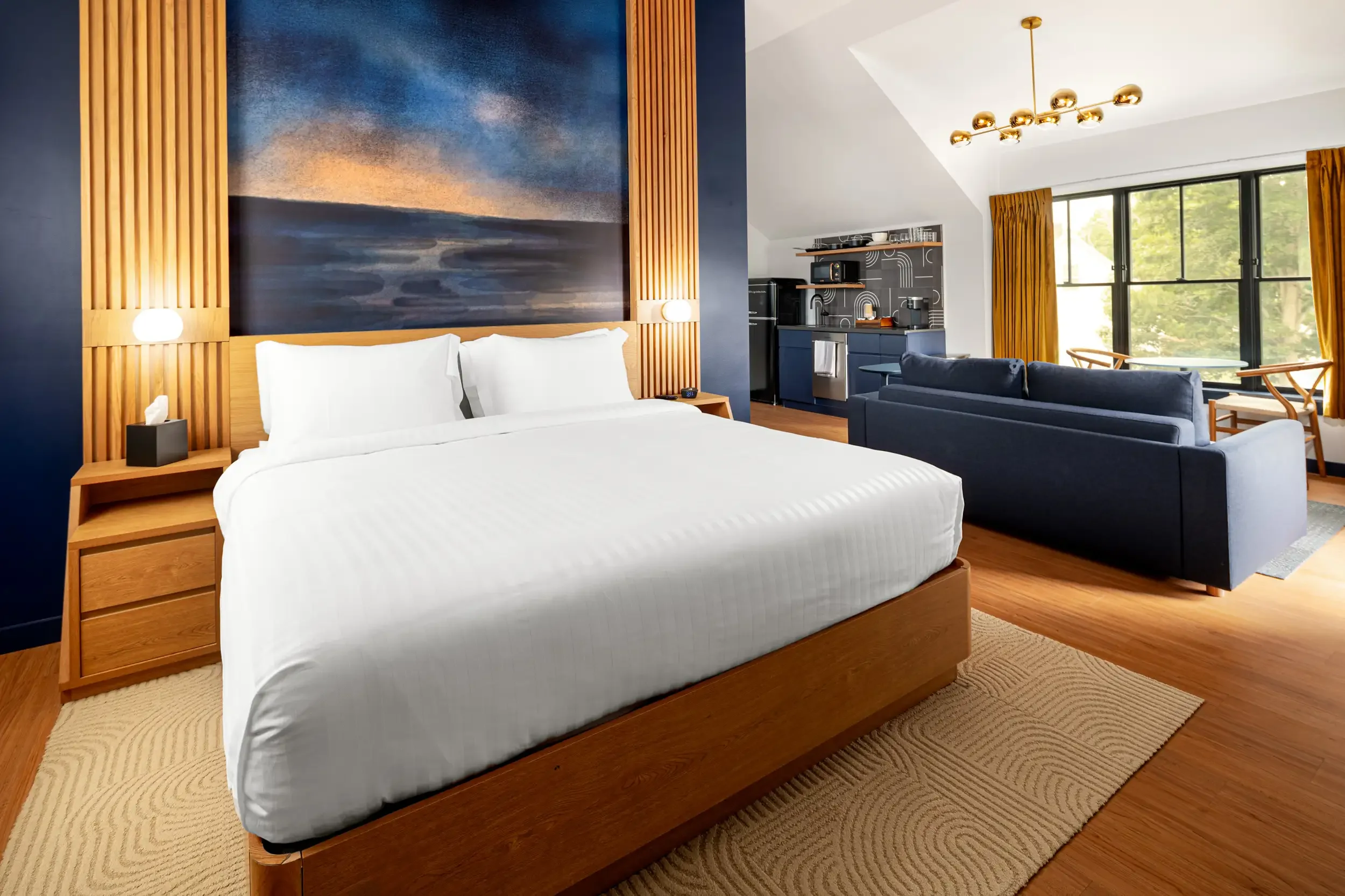 A modern hotel room with a large bed featuring white linens and a wooden headboard. There's a small nightstand on the left with a tissue box. The wall behind the bed has artistic blue and beige tones. To the right, there's a dark blue couch, a kitche