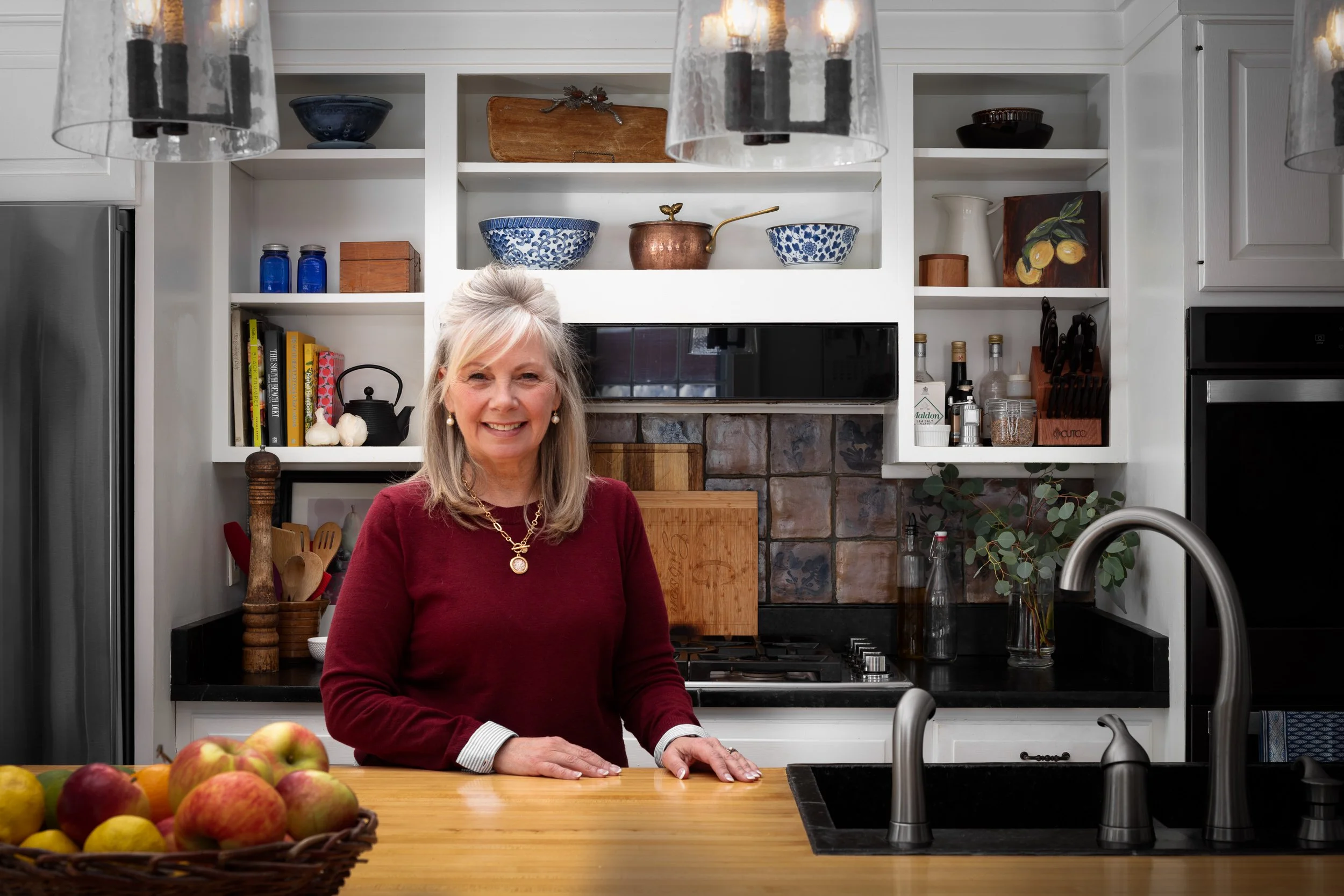 A woman with blonde hair and a red sweater smiling in a kitchen with white shelves, bowls, cutting boards, and a stovetop in the background. She is standing behind a wooden counter with a basket of apples and oranges, and a black sink with a silver f