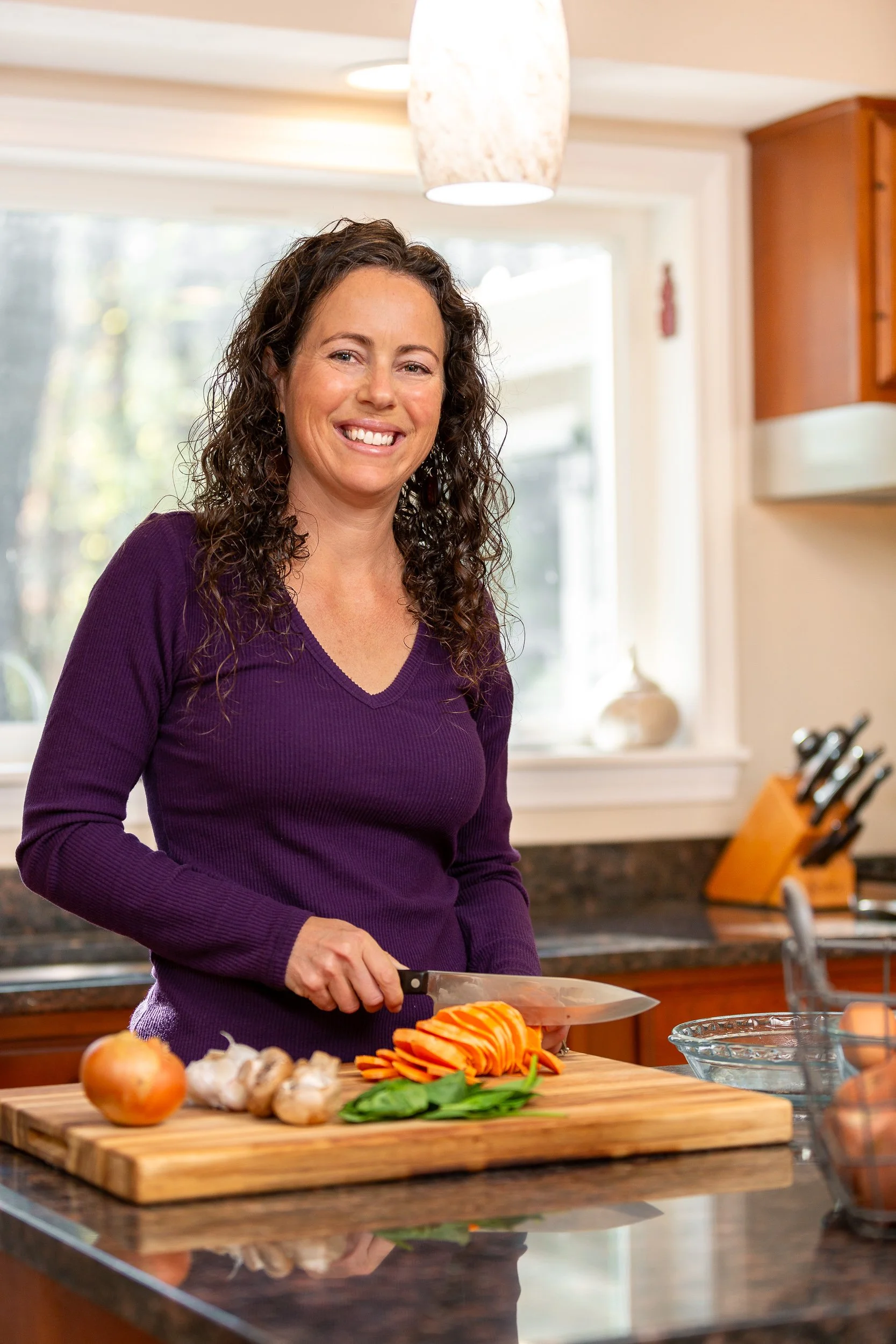A woman with curly hair smiling while chopping vegetables on a wooden cutting board in a kitchen.