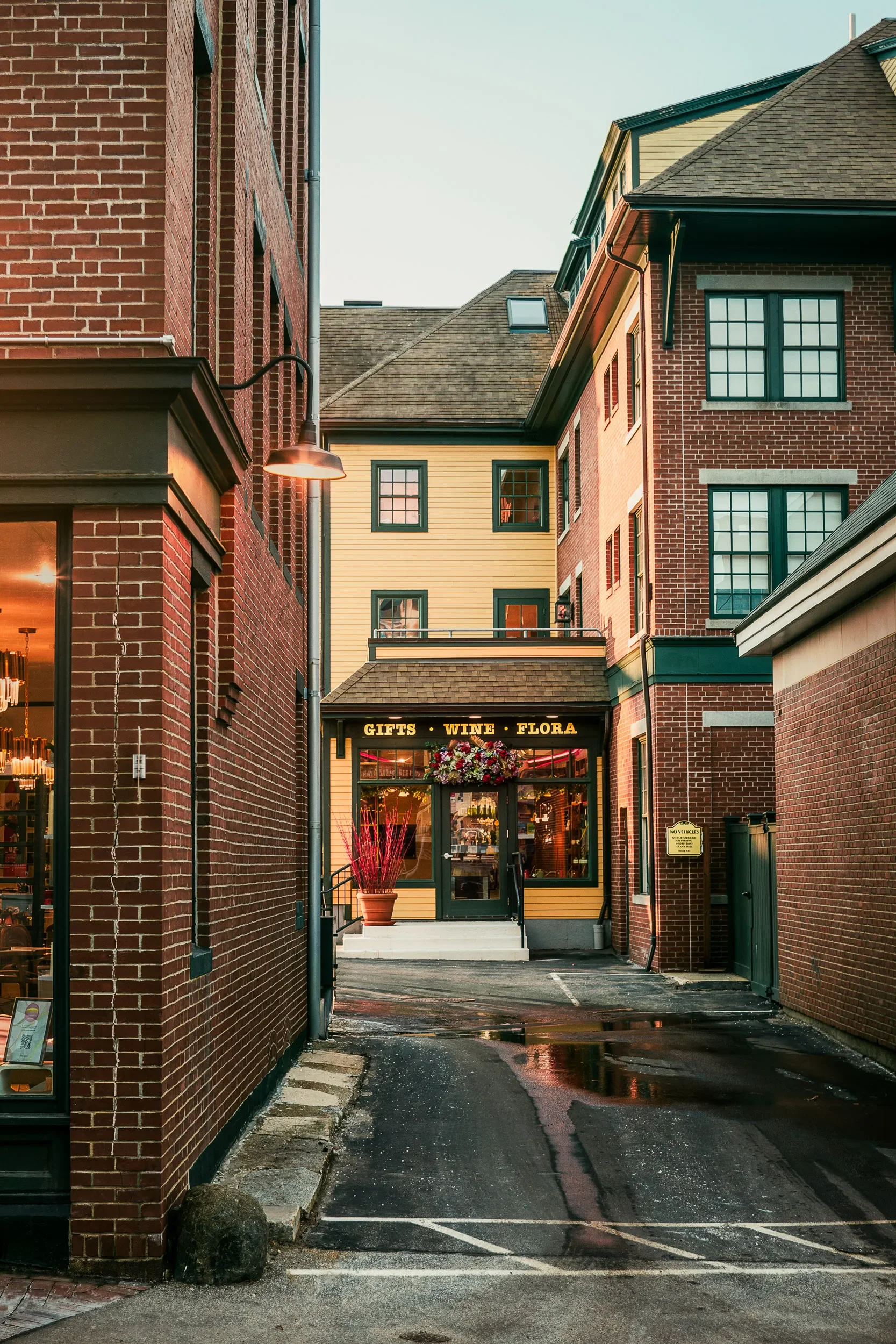 View of a small alleyway entrance to a retail store with a sign that reads gifts, wine, flora; the store is flanked by red brick buildings and a yellow building in the background, wet pavement reflecting the surroundings, and a potted plant with pink