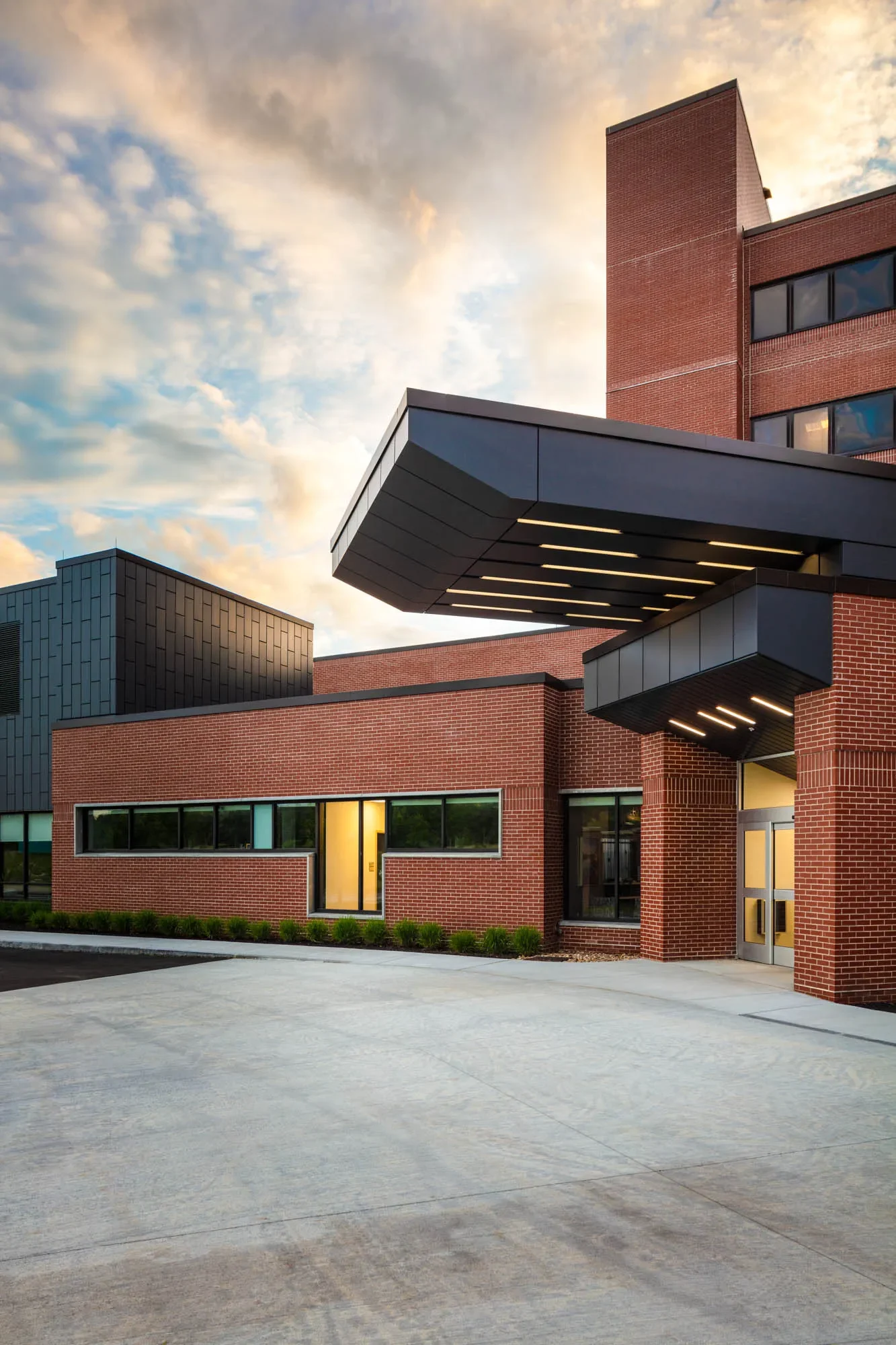 Modern building with red brick walls, large windows, and black metal accents, under a cloudy sky during sunset.