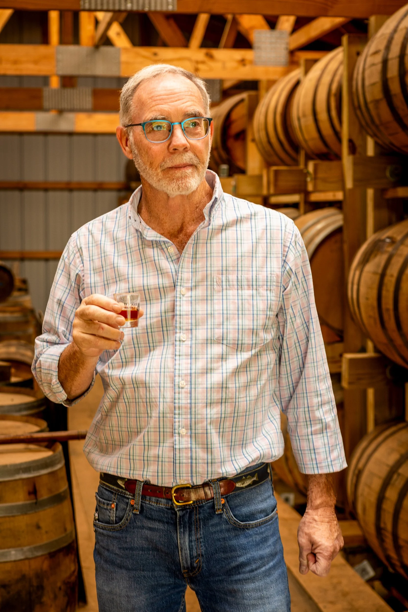 An older man with glasses and a beard standing in a cellar or warehouse with wooden barrels behind him, holding a small glass of dark liquor.