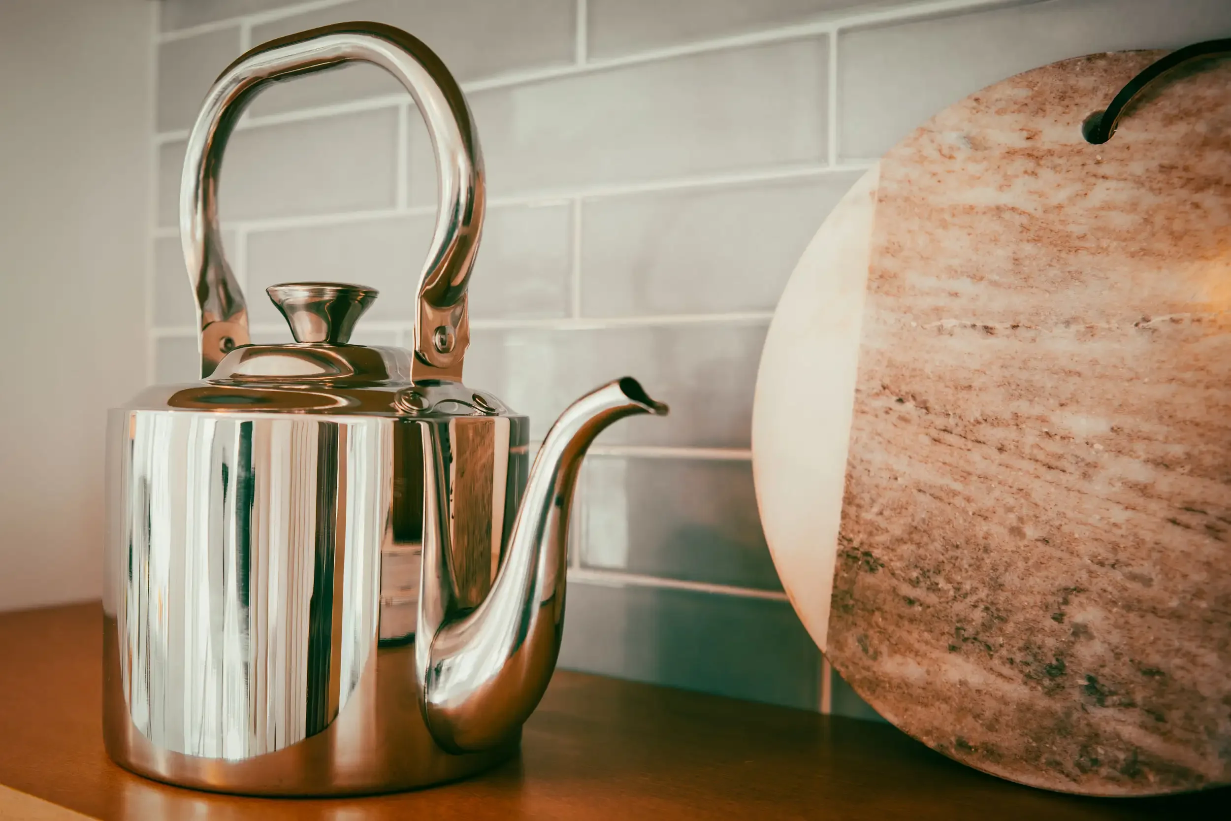 A shiny silver kettle with a curved handle and spout sitting on a wooden surface in front of a light-colored tiled wall and a portion of a brown granite countertop.