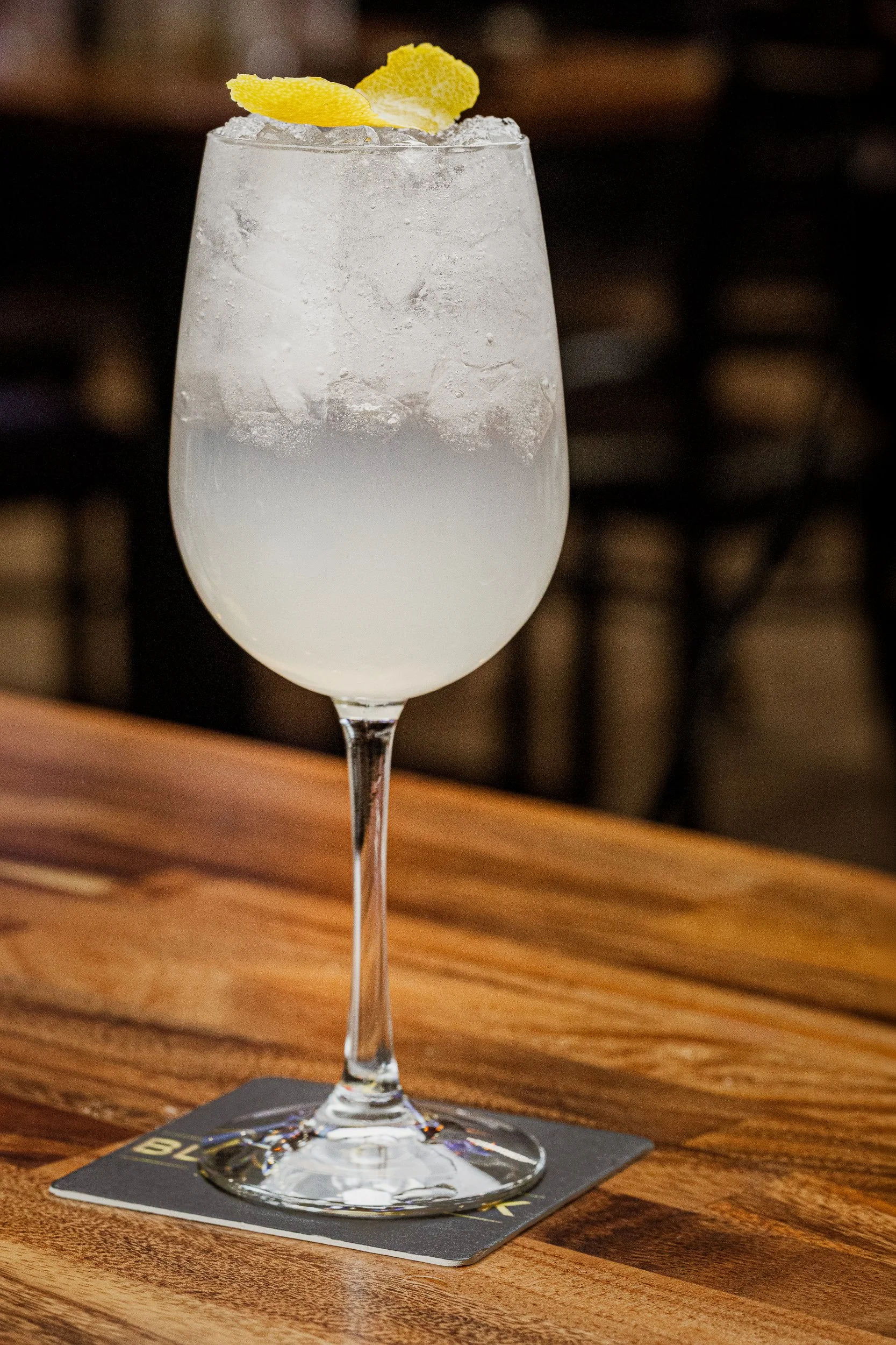 A cocktail glass filled with a clear, cloudy drink topped with lemon zest garnish, sitting on a wooden table.