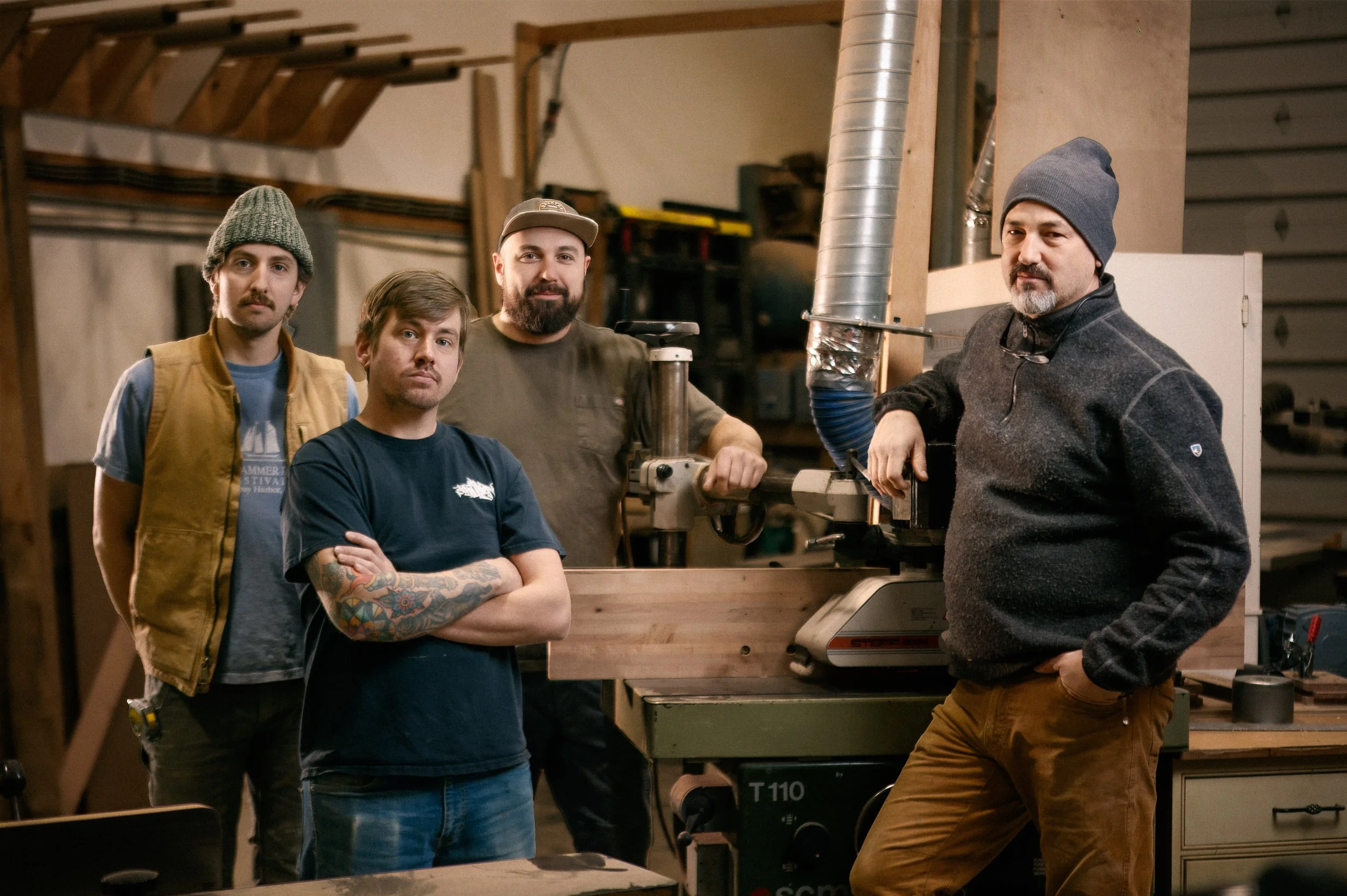 Four men in a woodworking shop, one using a machine and the others standing nearby, surrounded by woodworking tools and equipment.