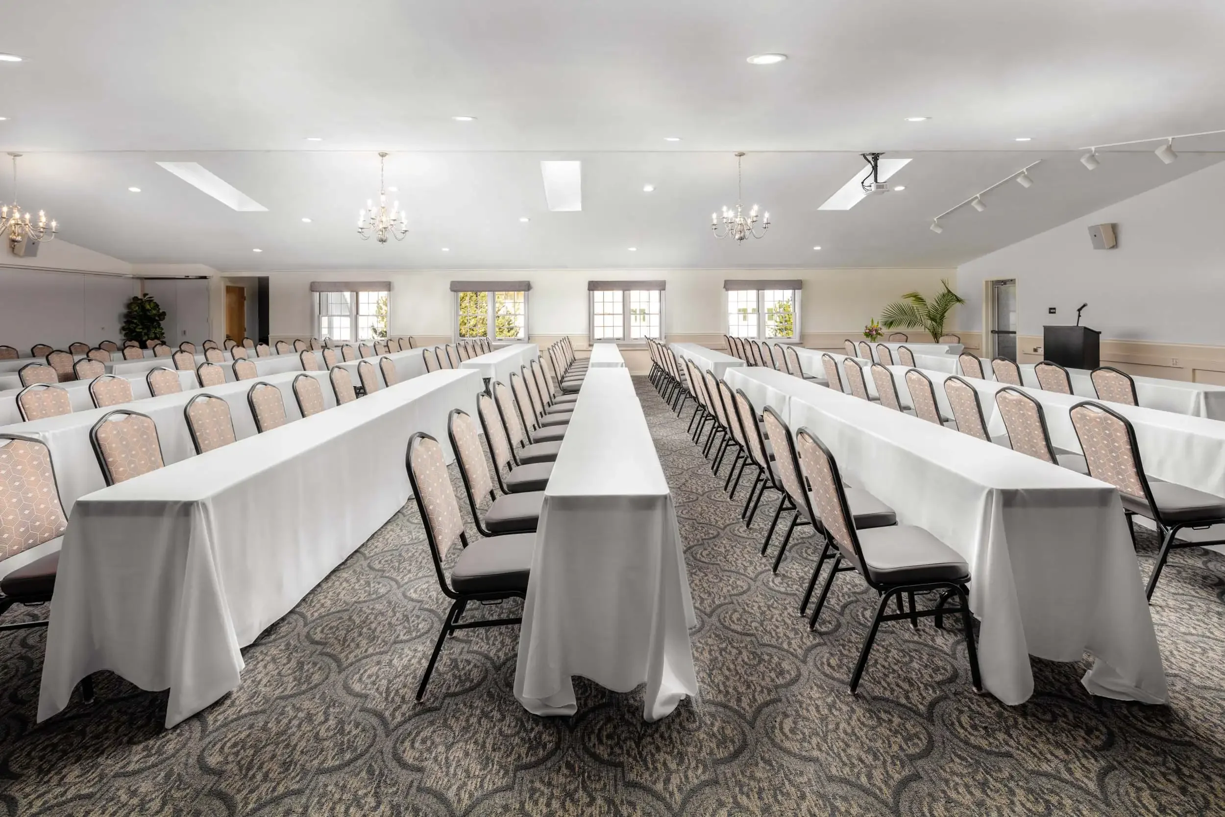 Empty conference room with long tables covered in white tablecloths, arranged in rows, and chairs with patterned upholstery. The room has windows, chandeliers, and a projector mounted on the ceiling.