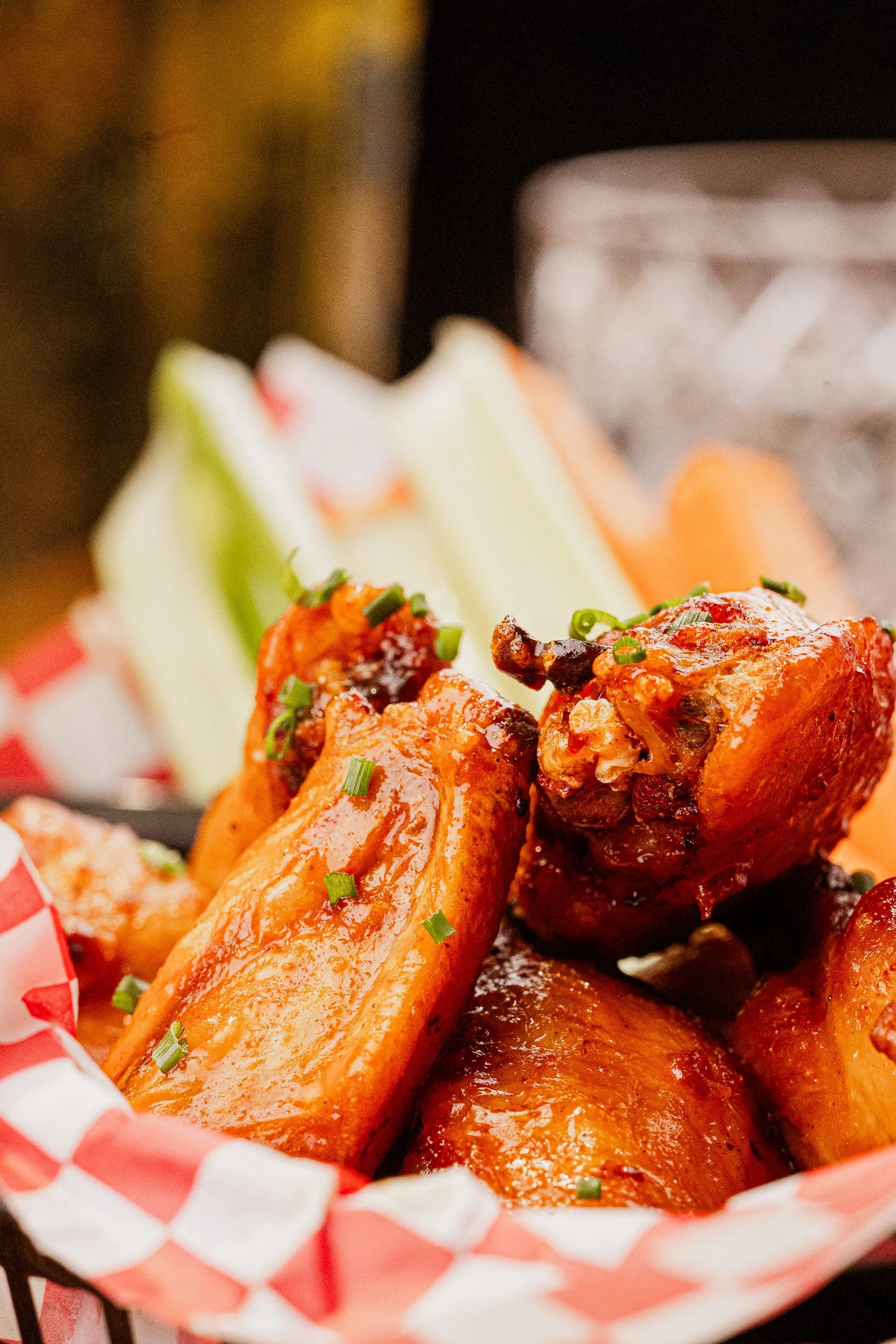 Close-up of buffalo chicken wings garnished with chopped green onions, served in a basket lined with red and white checkered paper, with celery and carrot sticks in the background.