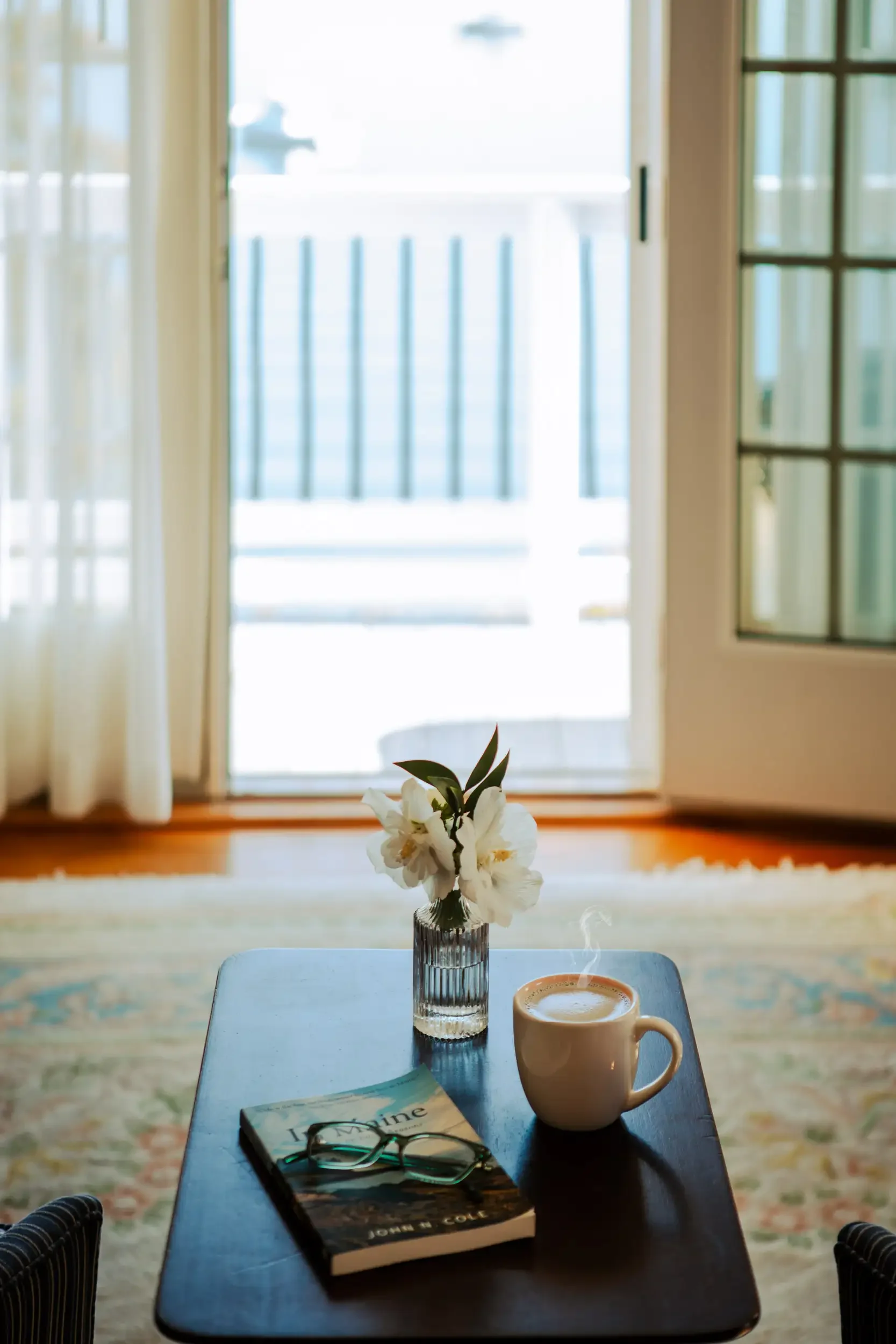 A table with a book, glasses, a vase with white flowers, a steaming cup, and a window with a balcony view in the background.