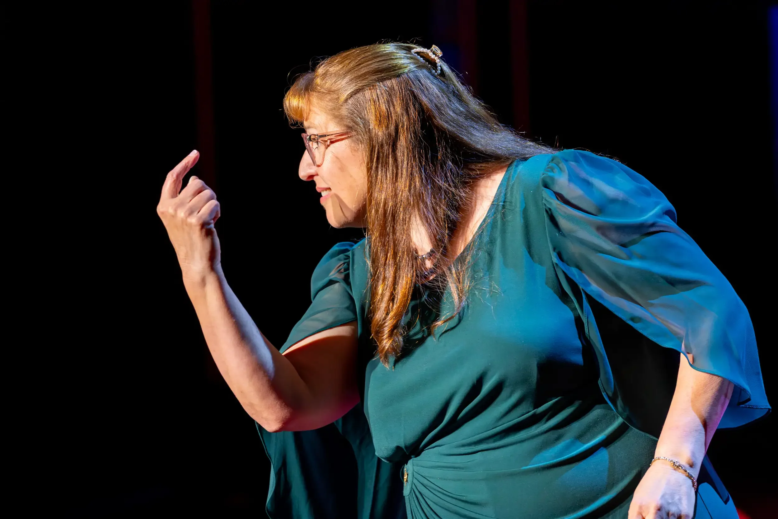 A woman with long brown hair, glasses, and a pearl hair accessory, wearing a teal blouse, making a thinking gesture with her hand.