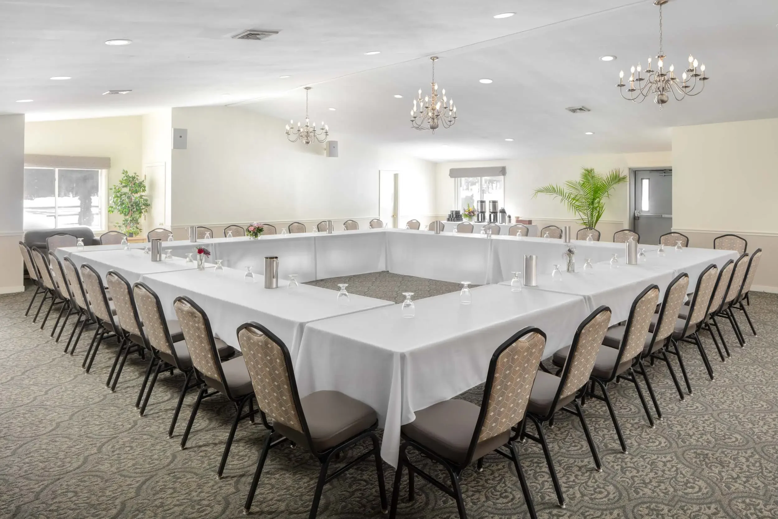 Empty banquet room with U-shaped table setup, decorated with small flower arrangements, surrounded by chairs, with chandeliers hanging from a white ceiling, windows on the walls, and a coffee station in the background.