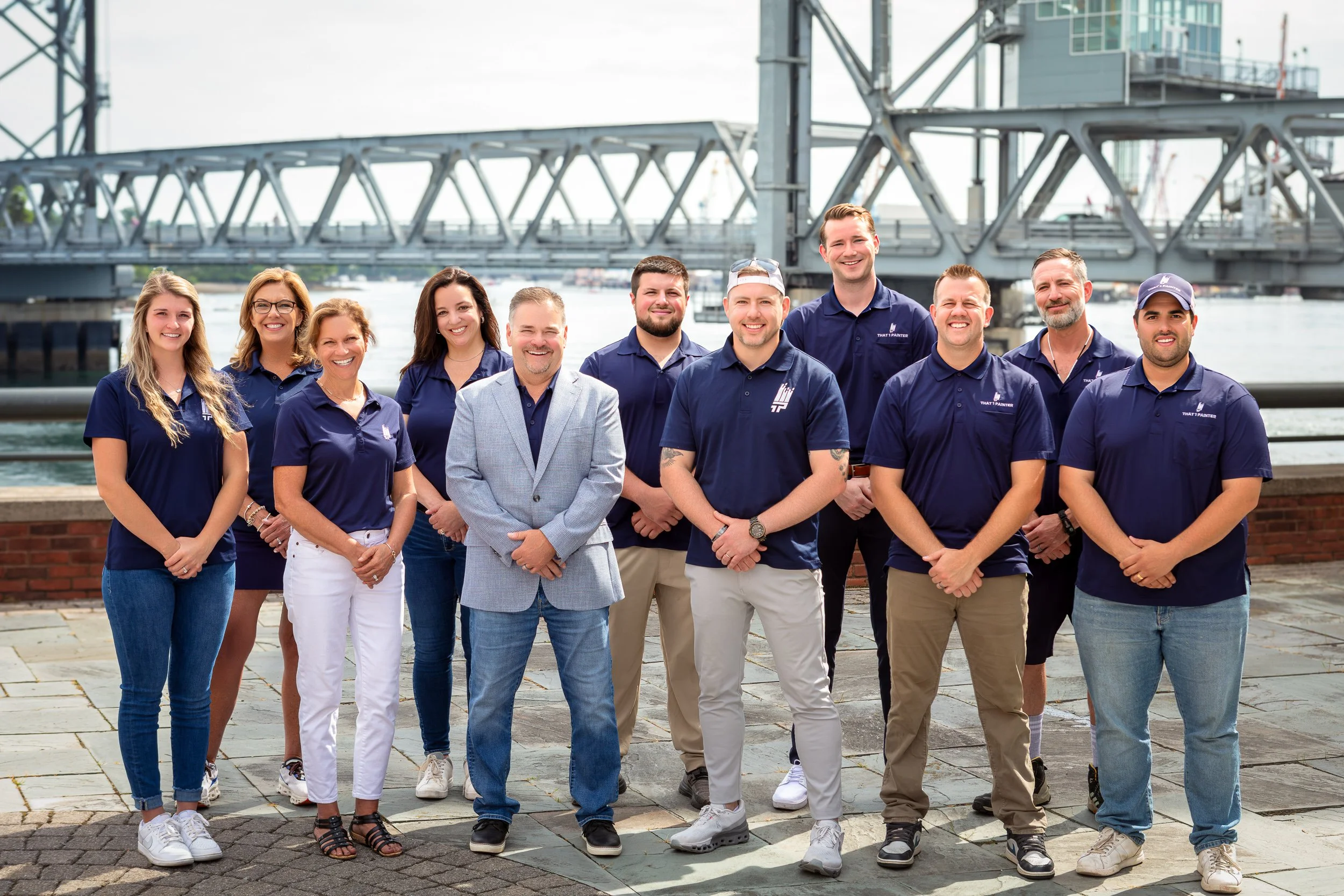 Group of twelve people standing outdoors under a bridge near water, smiling at the camera, dressed in navy blue shirts and casual pants.