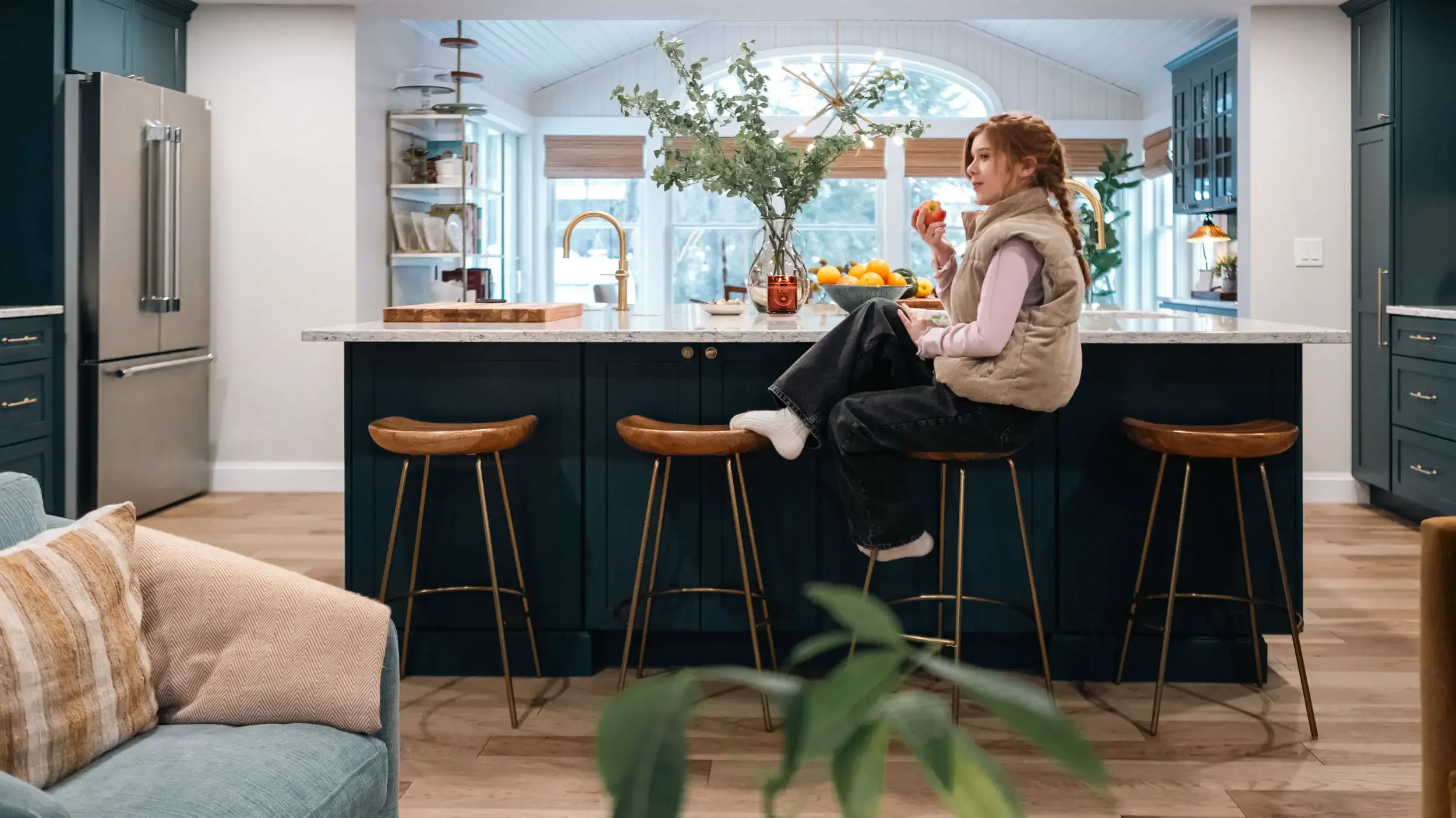 A young girl with red hair is sitting on a kitchen island, eating an apple, surrounded by a bowl of oranges and a vase with green foliage in a modern, well-lit kitchen.