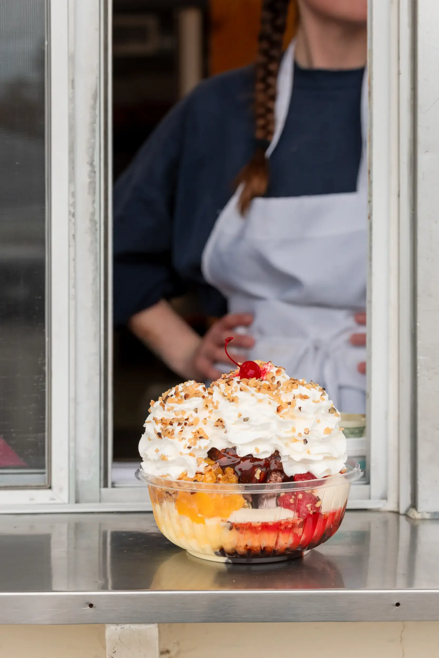 A bowl of ice cream sundae topped with whipped cream, crushed nuts, and a cherry, placed in front of a window with a person wearing a dark shirt and white apron looking out.