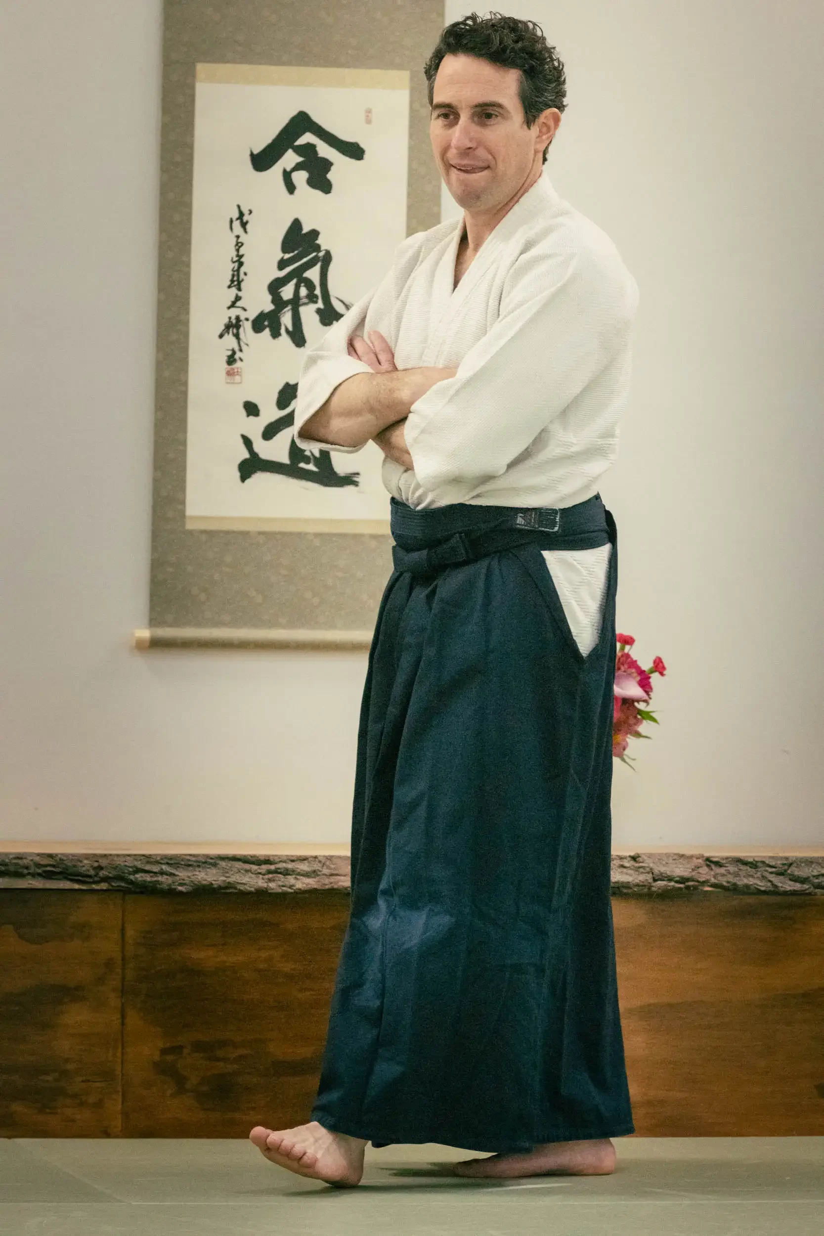 A person in traditional martial arts attire practicing a technique on a mat inside a dojo with Japanese calligraphy on a scroll in the background.