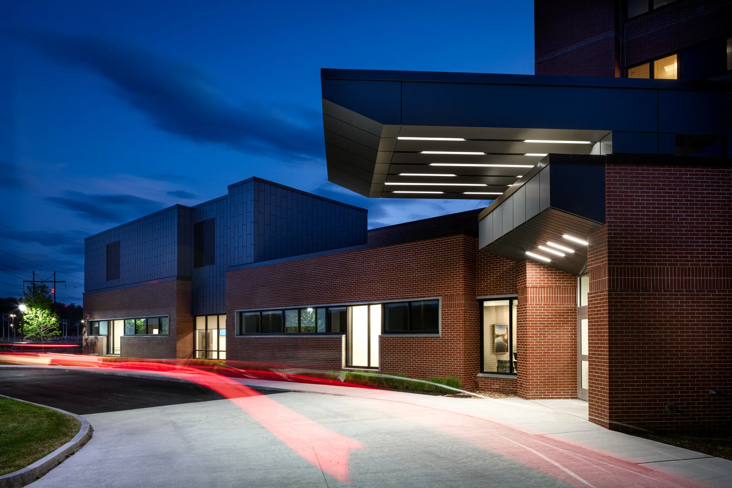 Modern building with brick and metal facade illuminated at night, with a curved driveway and streaks of red and white car light trails.