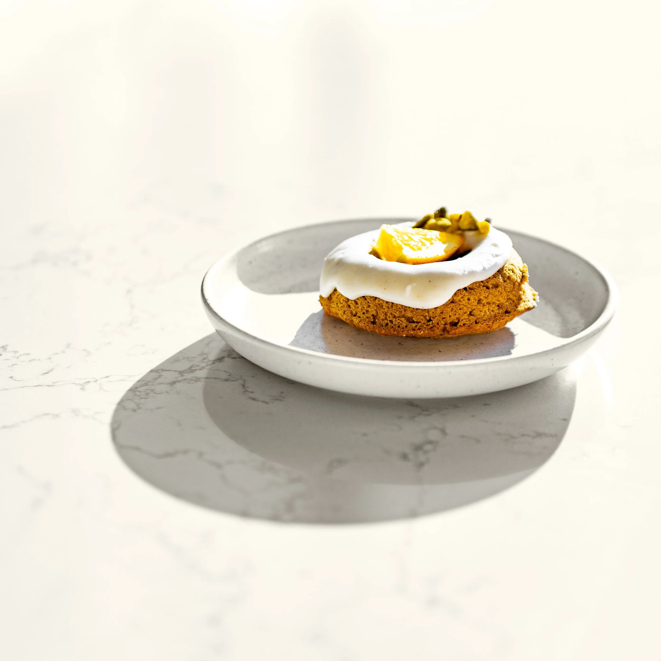 A cookie topped with white frosting, a lemon wedge, and chopped nuts on a white plate, with a marble surface and shadow.